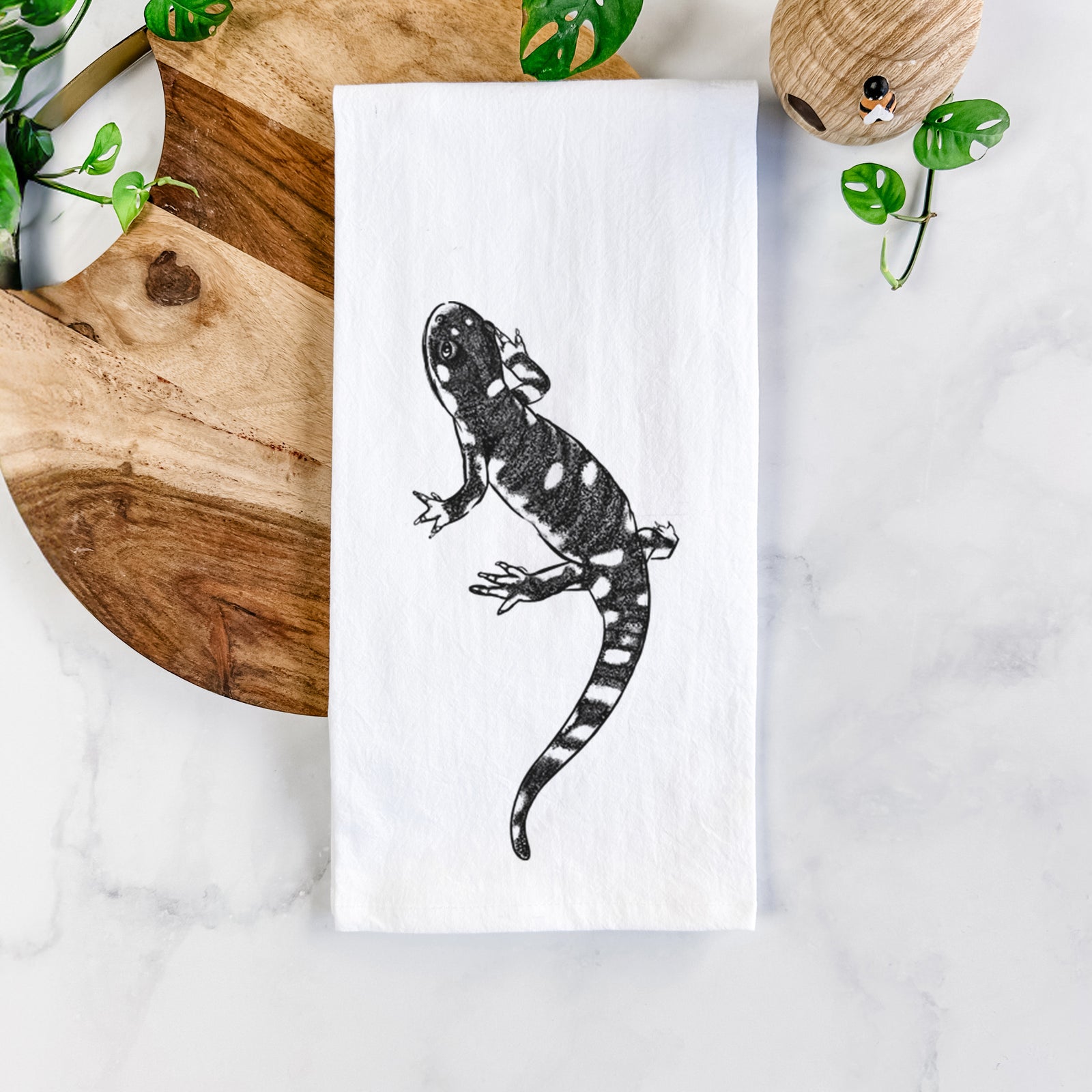 White cotton tea towel printed with a detailed black ink illustration of a spotted California tiger salamander, on a marble counter beside a round wooden cutting board and greenery.