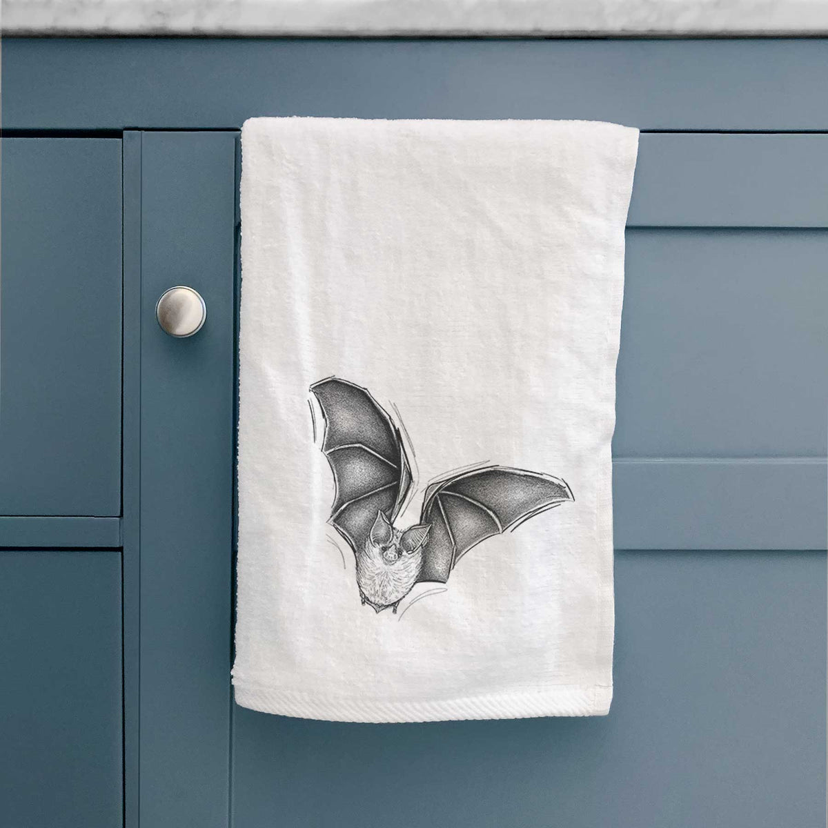 White hand towel printed with a detailed black ink illustration of a California leaf-nosed bat in flight, hanging on a cabinet door.