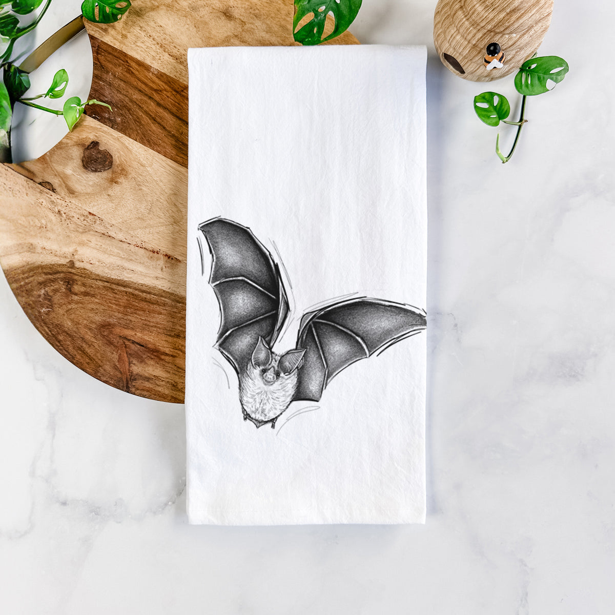 White cotton tea towel printed with a detailed black ink illustration of a California leaf-nosed bat in flight, on a marble counter beside a round wooden cutting board and greenery.