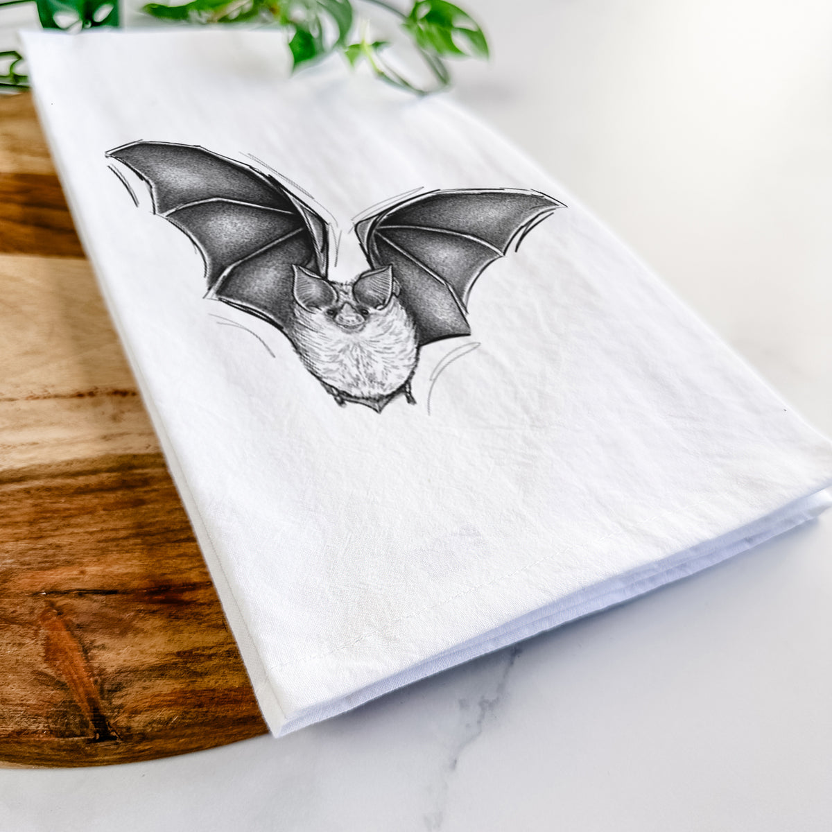 Close-up of a folded white tea towel on a wooden board, showing a detailed black ink illustration of a California leaf-nosed bat in flight.