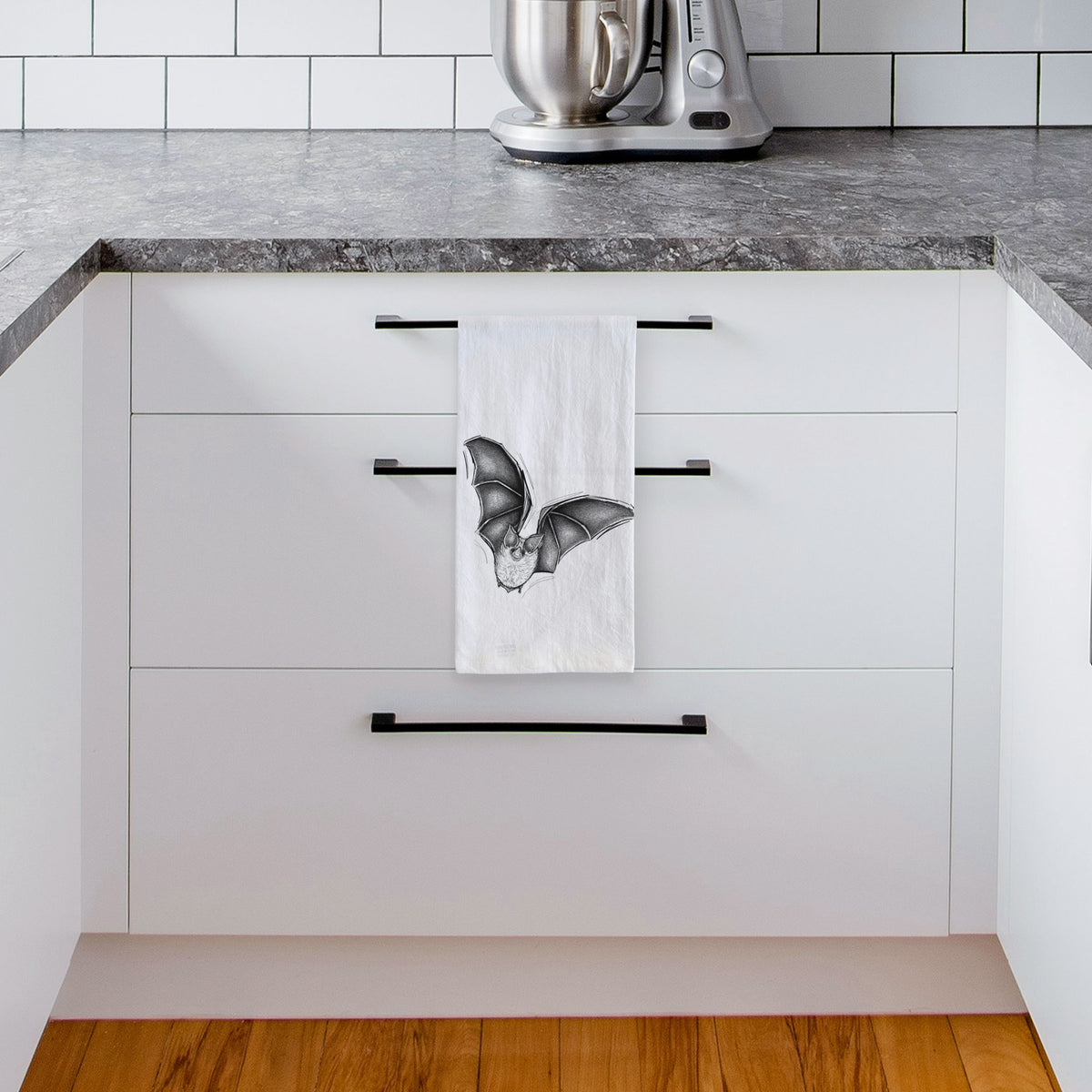 White tea towel printed with a detailed black ink illustration of a California leaf-nosed bat in flight, hanging from a drawer handle in a bright white kitchen.