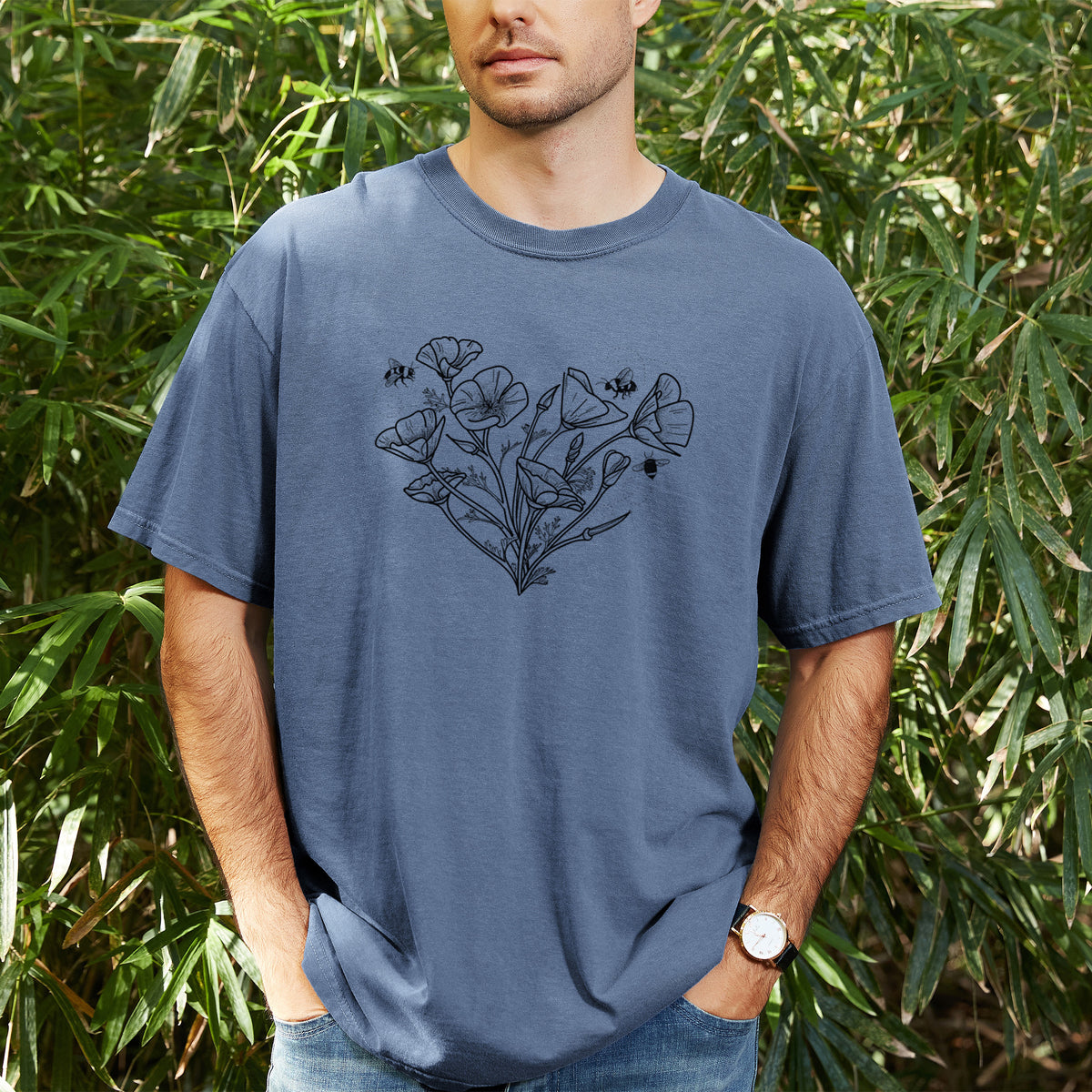 Man wearing a Comfort Colors t-shirt printed with a black ink line drawing of California poppies arranged in a heart shape, with small bees, outdoors.