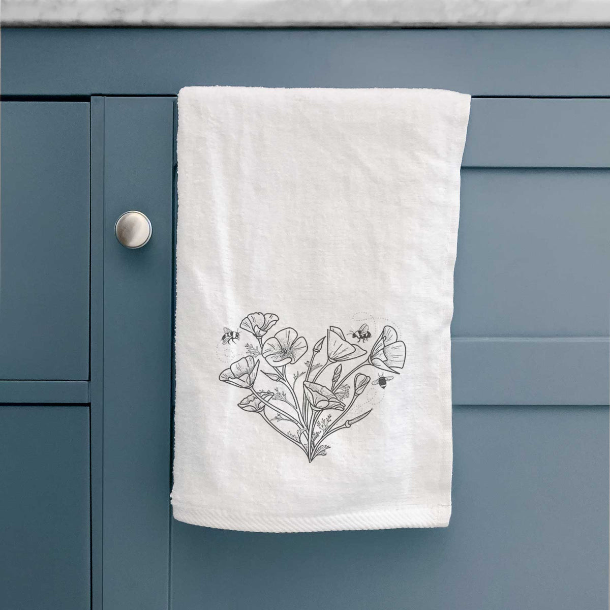 White hand towel printed with a black ink line drawing of California poppies arranged in a heart shape, with small bees, hanging on a cabinet door.