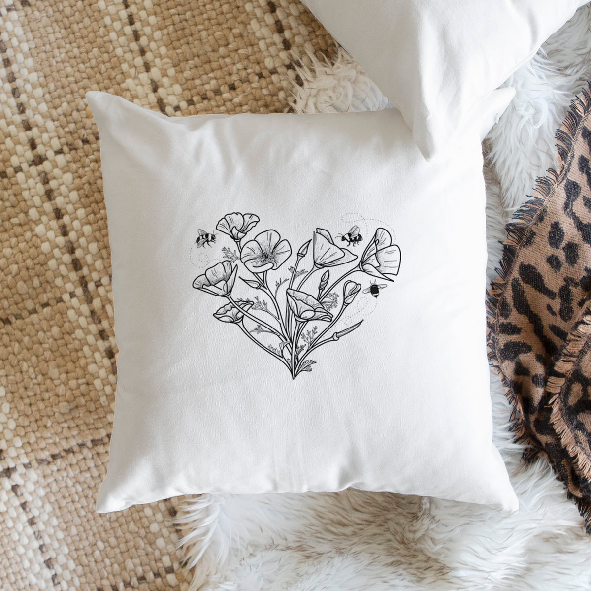 White pillow cover printed with a black ink line drawing of California poppies arranged in a heart shape, with small bees, on a woven chair.