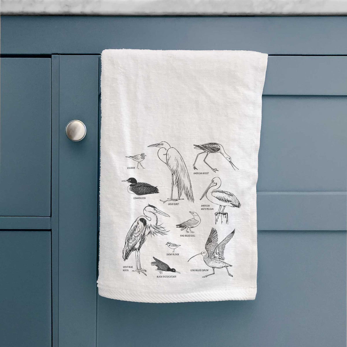 White hand towel printed with a chart of black ink California shorebirds, including herons, egrets, avocets and sandpipers, hanging on a cabinet door.