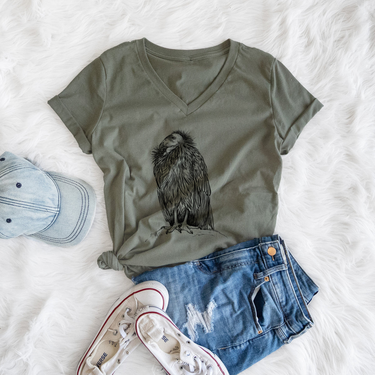 Military green women's relaxed fit cotton V-neck t-shirt printed with a detailed black ink illustration of a California condor perched with folded wings, styled flat on a white faux fur rug with a cap, jeans and sneakers.