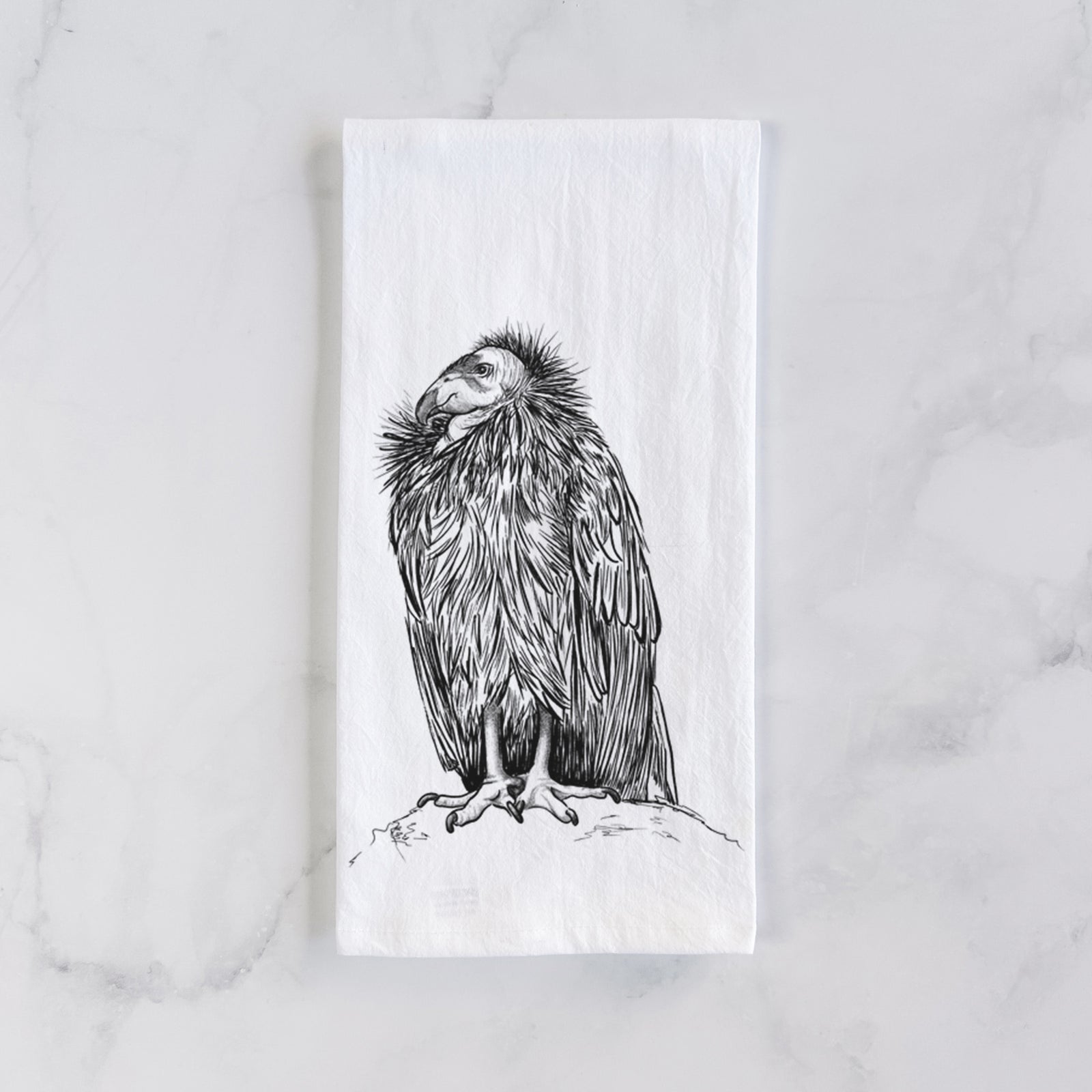 White cotton tea towel printed with a detailed black ink illustration of a California condor perched with folded wings, on a marble counter beside a round wooden cutting board and greenery.