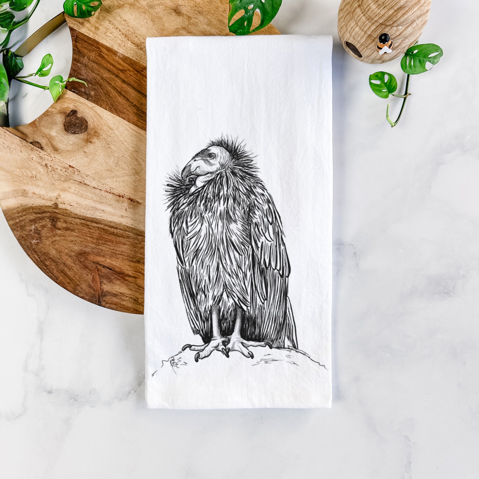 White cotton tea towel printed with a detailed black ink illustration of a California condor perched with folded wings, on a marble counter beside a round wooden cutting board and greenery.