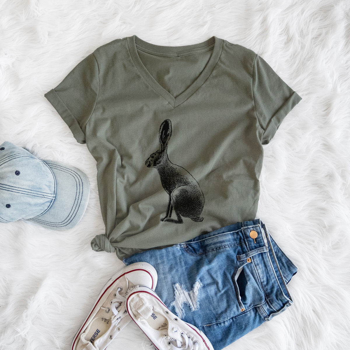 Military green women's relaxed fit cotton V-neck t-shirt printed with a detailed black ink illustration of a black-tailed jackrabbit sitting in profile with tall ears, styled flat on a white faux fur rug with a cap, jeans and sneakers.