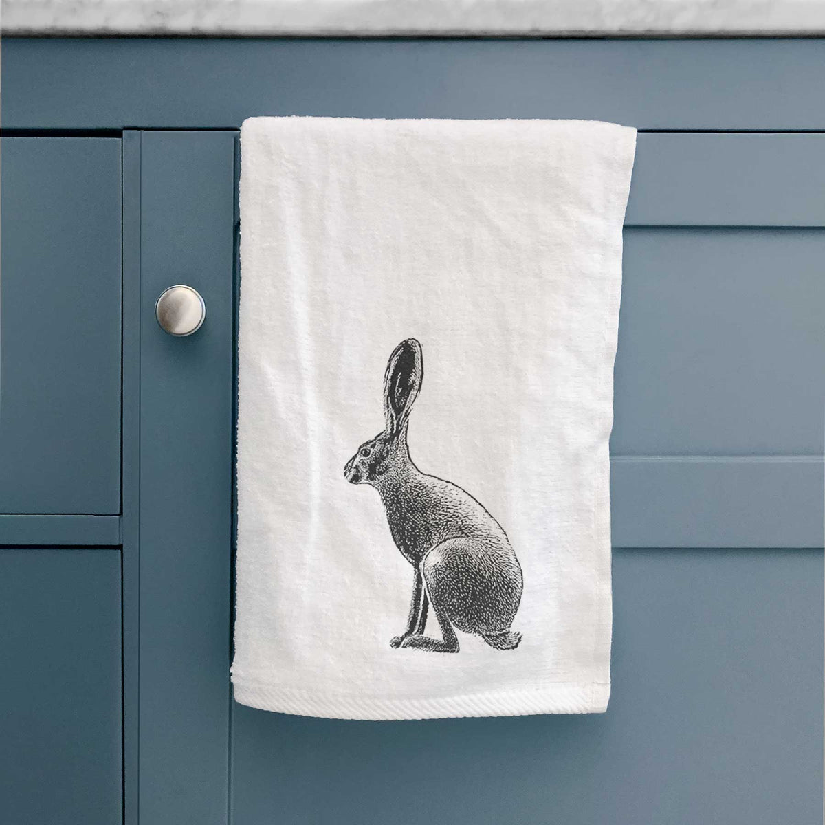 White hand towel printed with a detailed black ink illustration of a black-tailed jackrabbit sitting in profile with tall ears, hanging on a cabinet door.