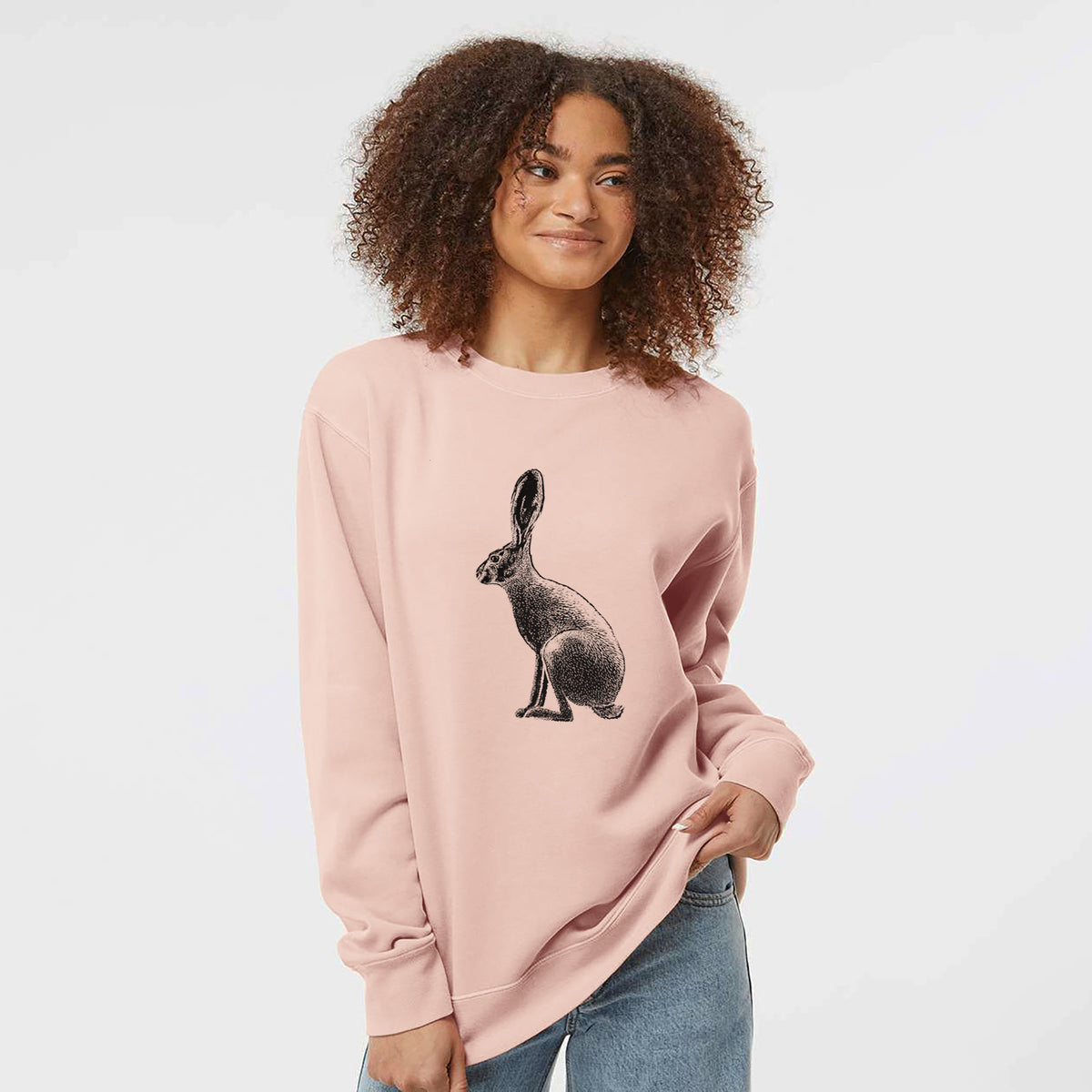 Model wearing a dusty pink pigment dyed crewneck sweatshirt printed with a detailed black ink illustration of a black-tailed jackrabbit sitting in profile with tall ears.