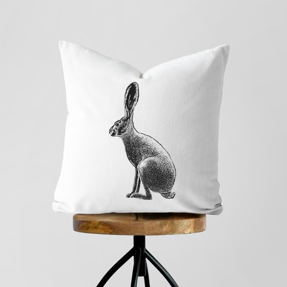 White pillow cover printed with a detailed black ink illustration of a black-tailed jackrabbit sitting in profile with tall ears, on a wooden stool.