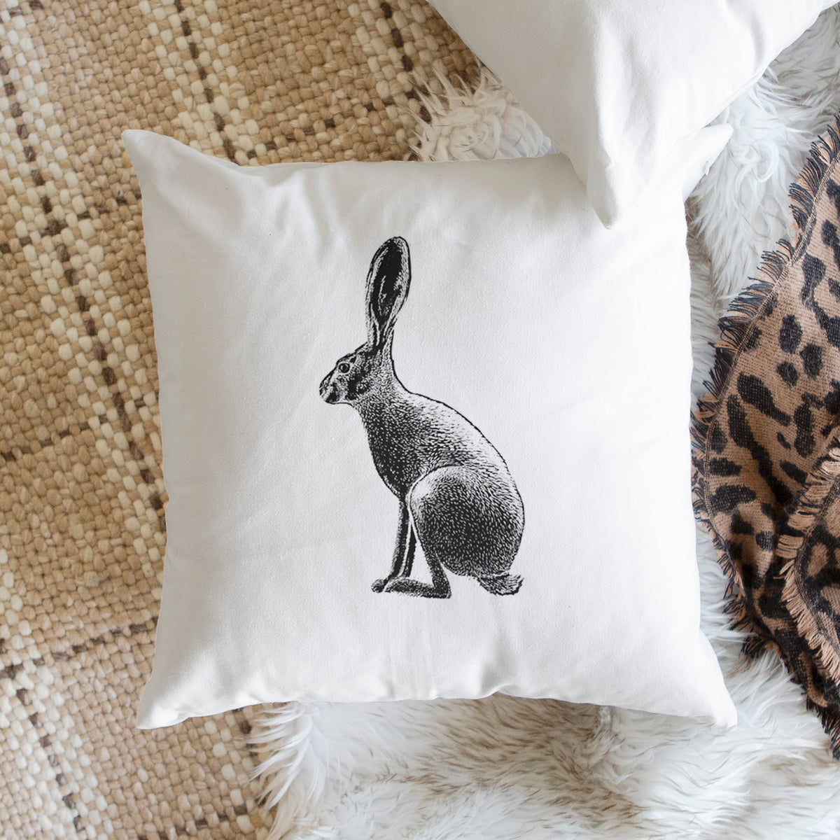 White pillow cover printed with a detailed black ink illustration of a black-tailed jackrabbit sitting in profile with tall ears, on a woven chair.