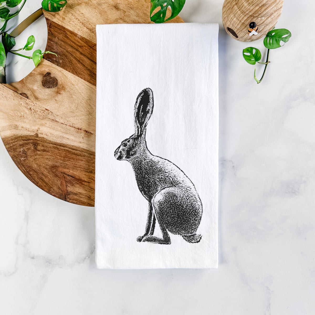 White cotton tea towel printed with a detailed black ink illustration of a black-tailed jackrabbit sitting in profile with tall ears, on a marble counter beside a round wooden cutting board and greenery.
