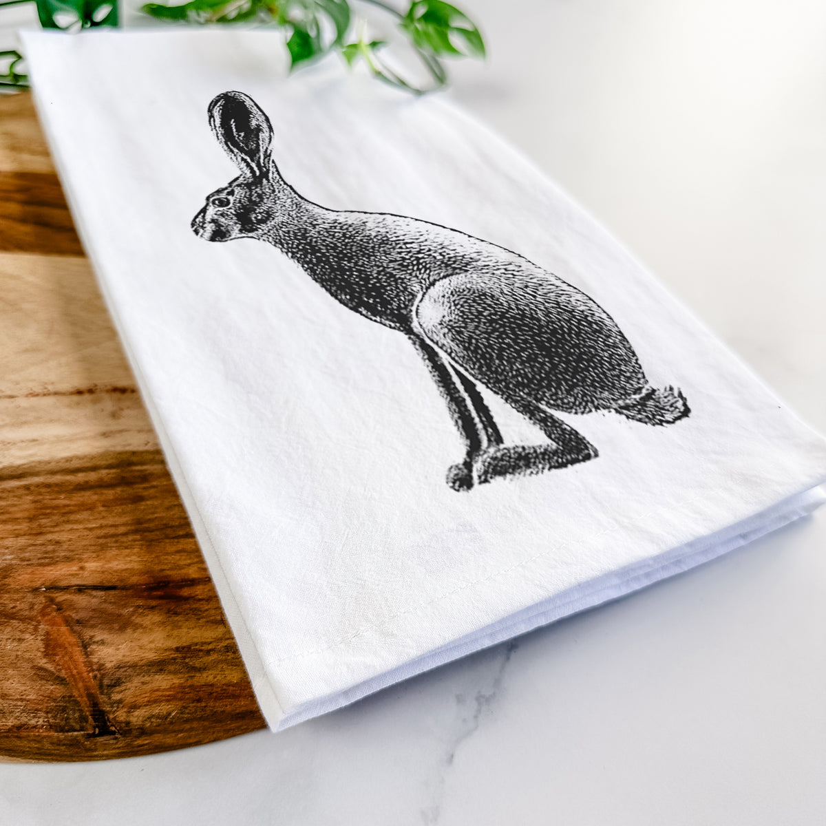 Close-up of a folded white tea towel on a wooden board, showing a detailed black ink illustration of a black-tailed jackrabbit sitting in profile with tall ears.