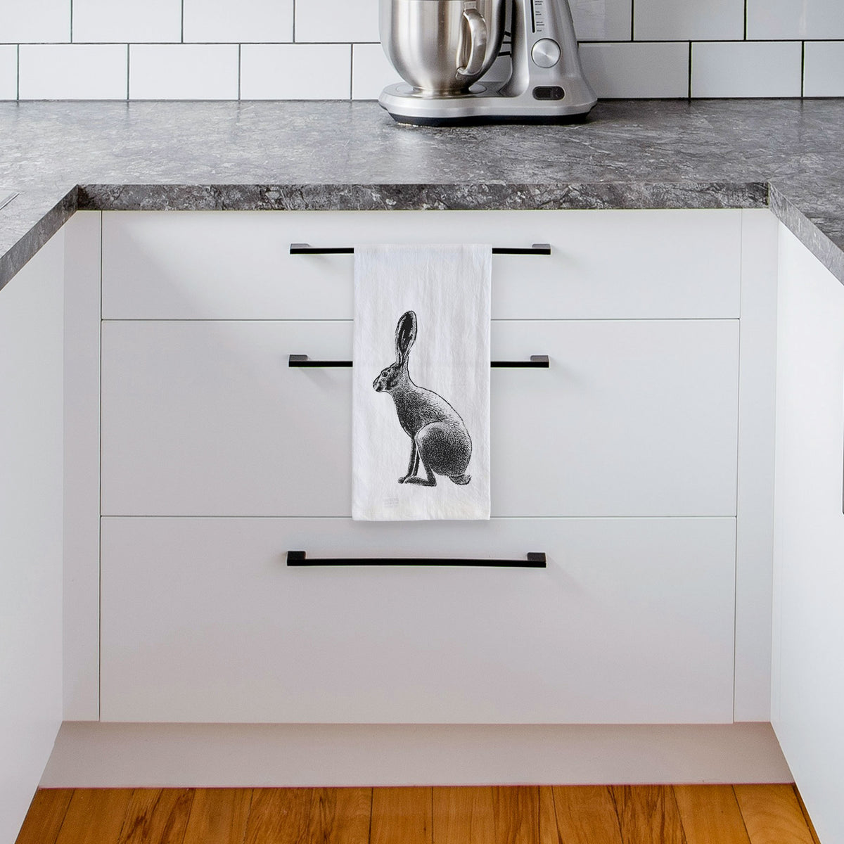 White tea towel printed with a detailed black ink illustration of a black-tailed jackrabbit sitting in profile with tall ears, hanging from a drawer handle in a bright white kitchen.