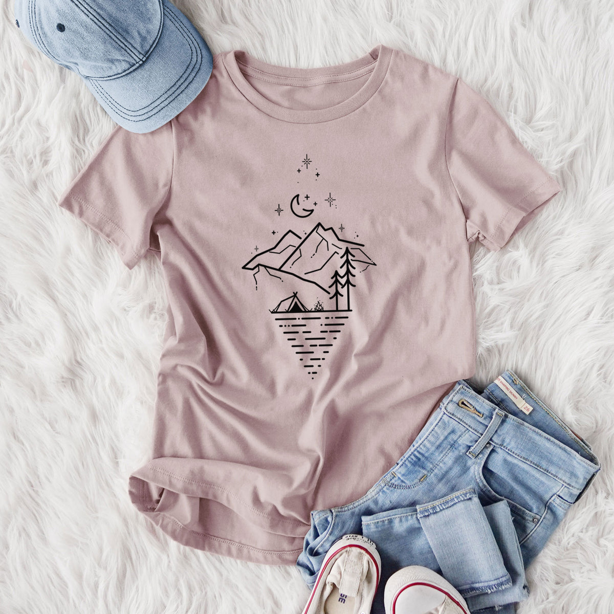 Women's relaxed fit cotton t-shirt printed with black line art of mountains, pine trees, a crescent moon and stars above a lake inside a diamond outline, styled flat on a white faux fur rug with jeans.