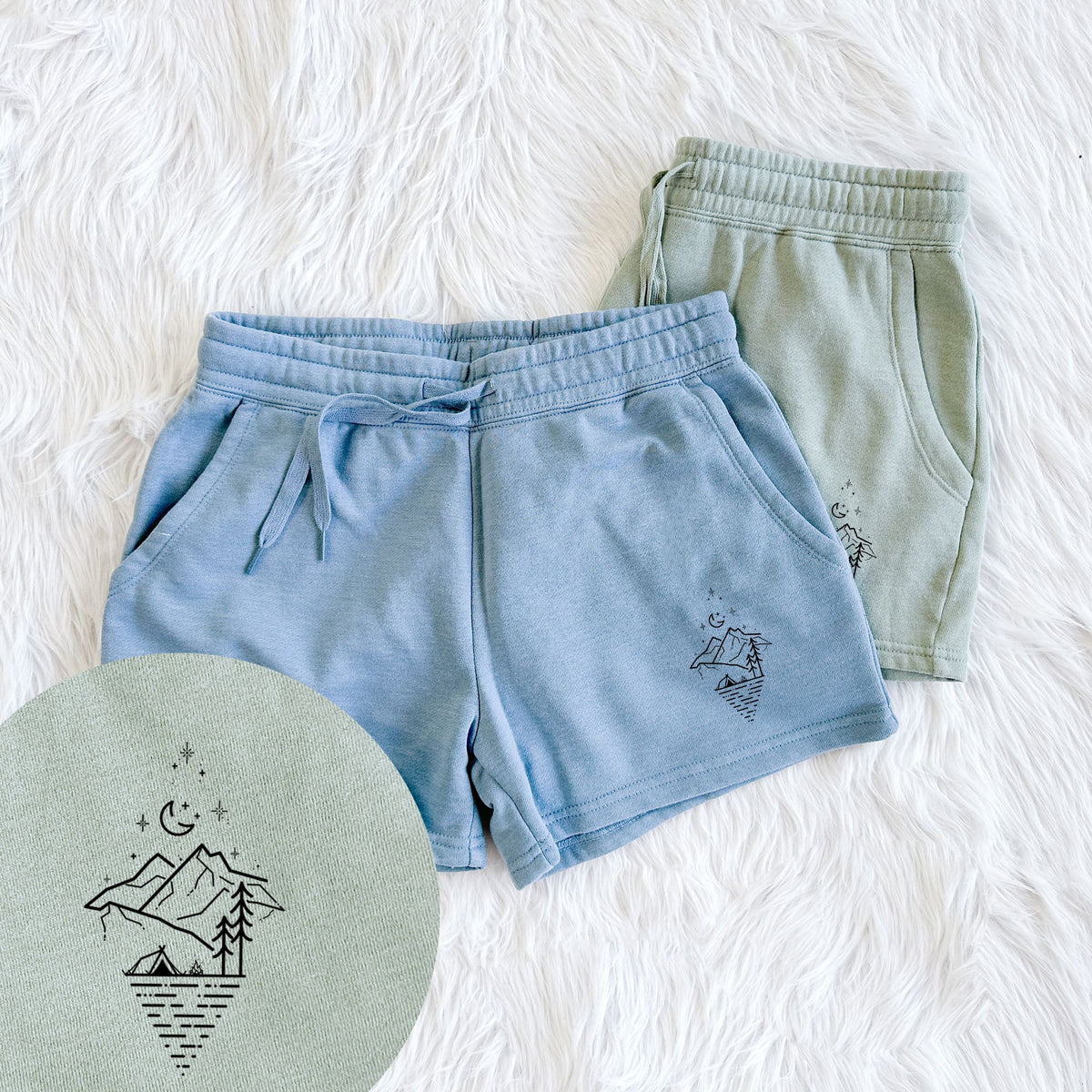Two pairs of Cali Wave shorts in different colors printed with black line art of mountains, pine trees, a crescent moon and stars above a lake inside a diamond outline, with a close-up of the print.