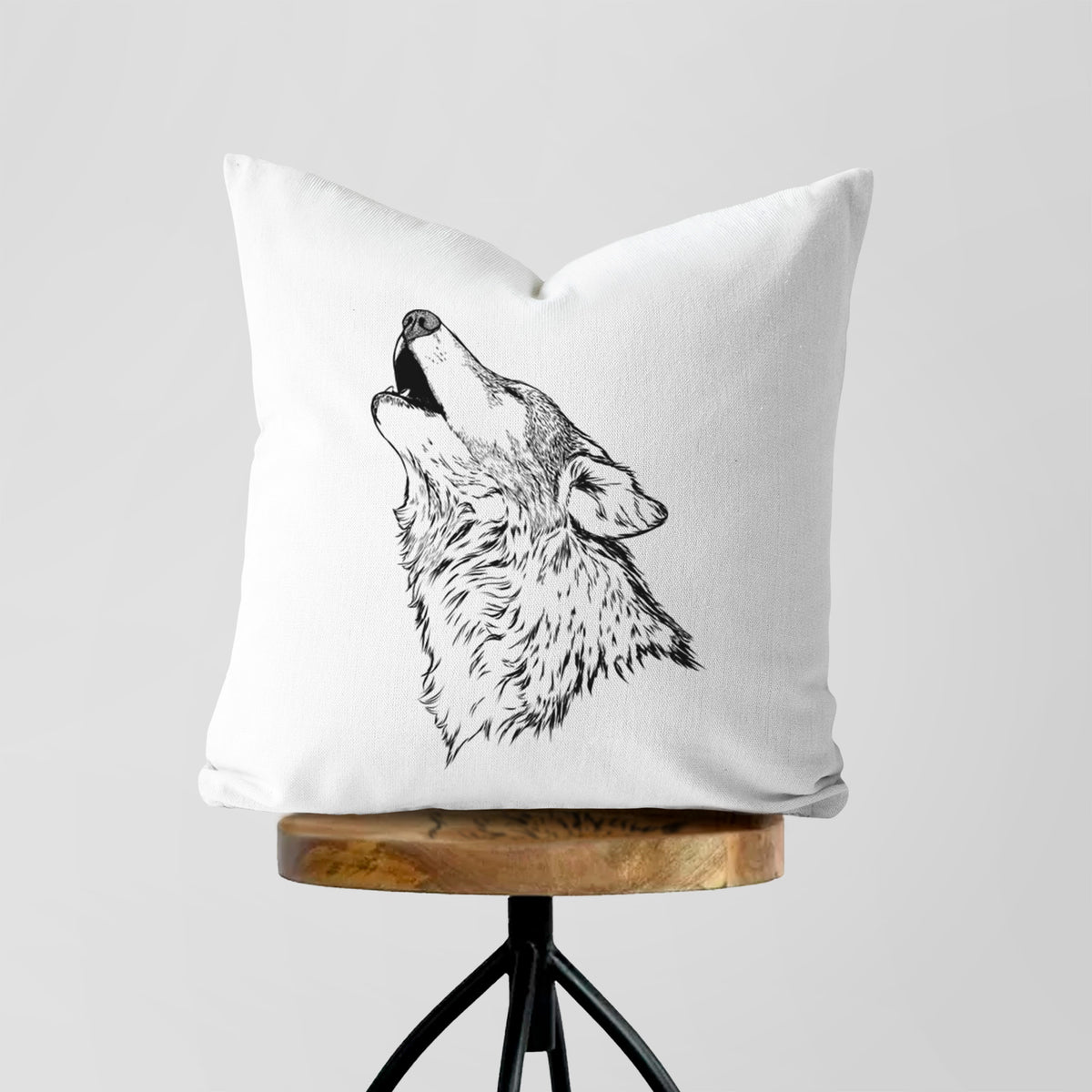 White pillow cover printed with a detailed black ink illustration of a grey wolf's head and shoulders, howling upward, on a wooden stool.