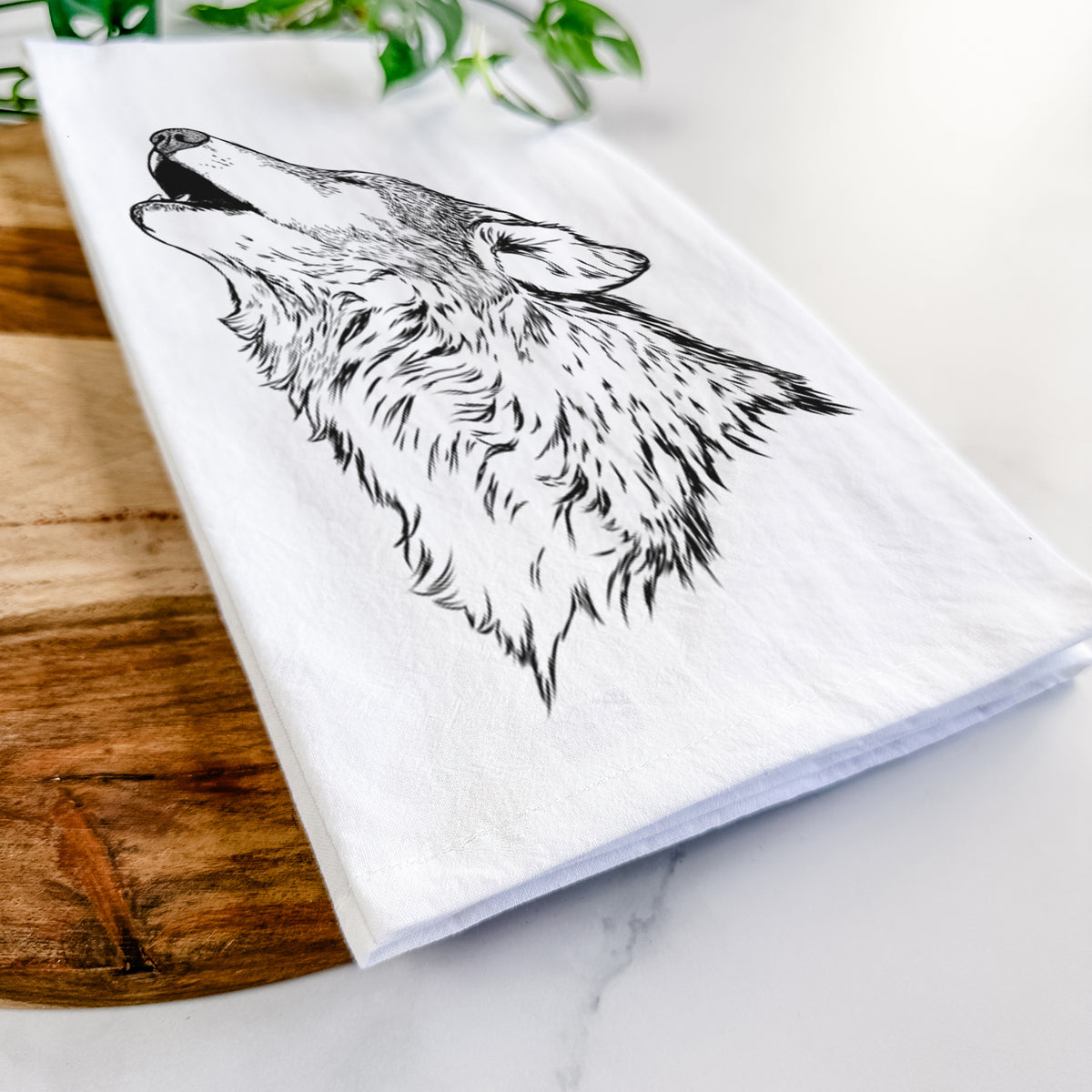 Close-up of a folded white tea towel on a wooden board, showing a detailed black ink illustration of a grey wolf's head and shoulders, howling upward.
