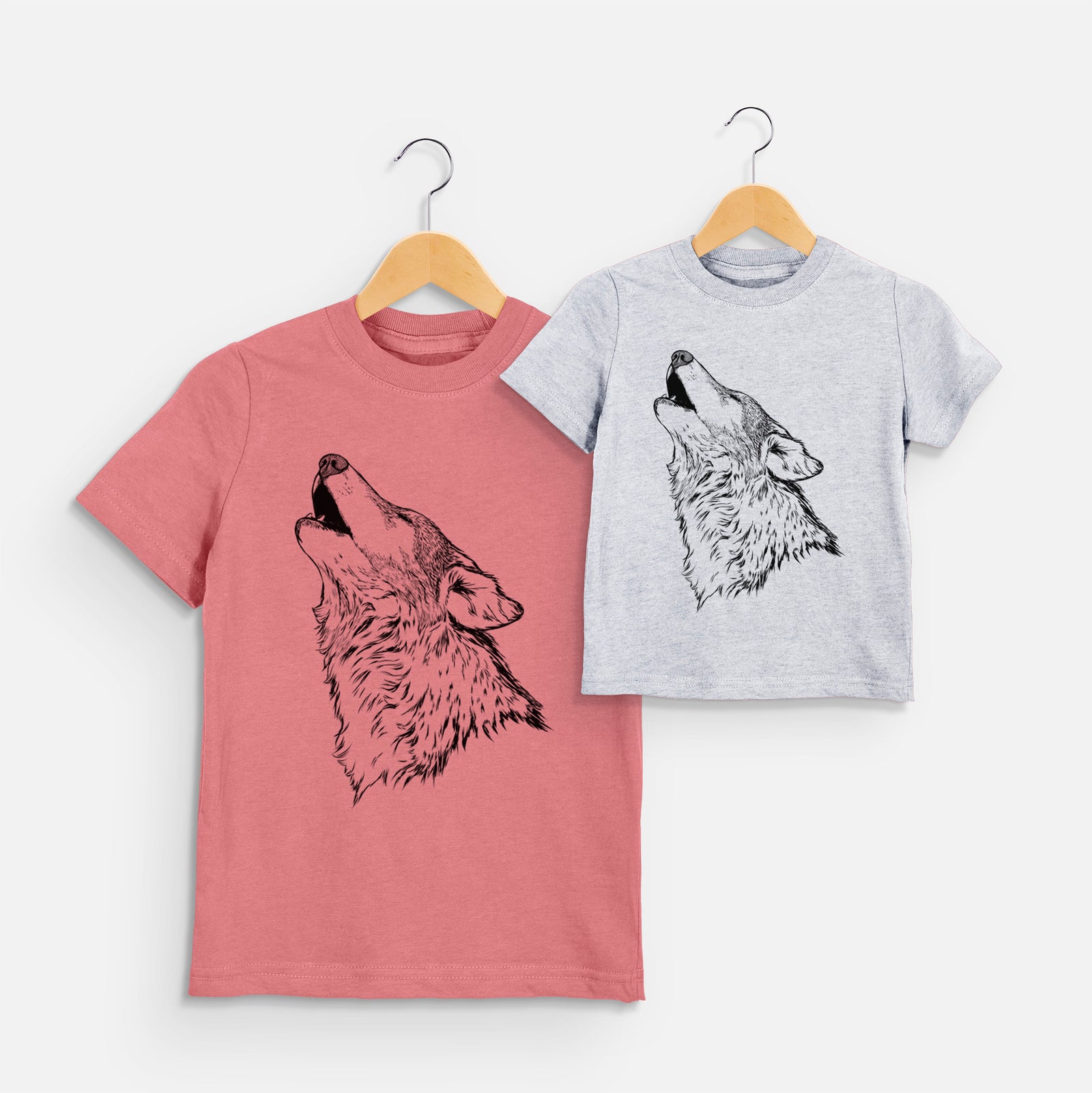 Two youth t-shirts in different colors on hangers, printed with a detailed black ink illustration of a grey wolf's head and shoulders, howling upward.