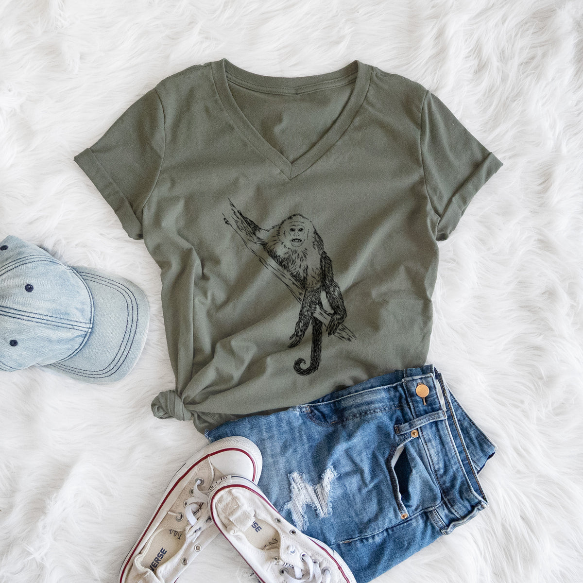 Military green women's relaxed fit cotton V-neck t-shirt printed with a detailed black ink illustration of a capuchin monkey climbing a branch, styled flat on a white faux fur rug with a cap, jeans and sneakers.
