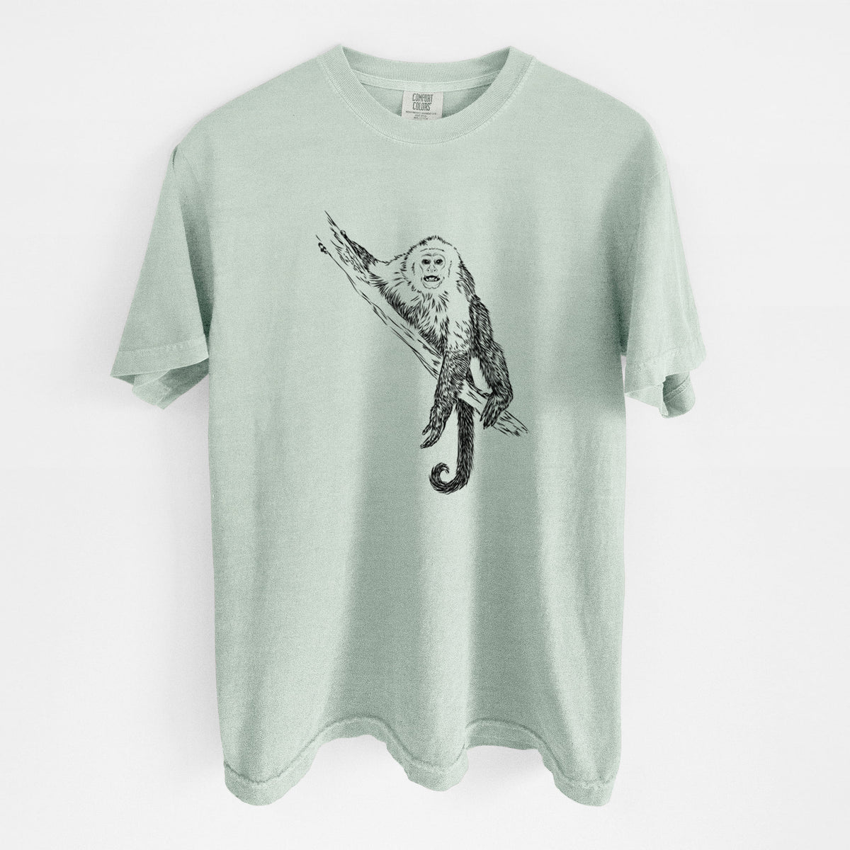 Bay green Comfort Colors t-shirt laid flat, printed with a detailed black ink illustration of a capuchin monkey climbing a branch.