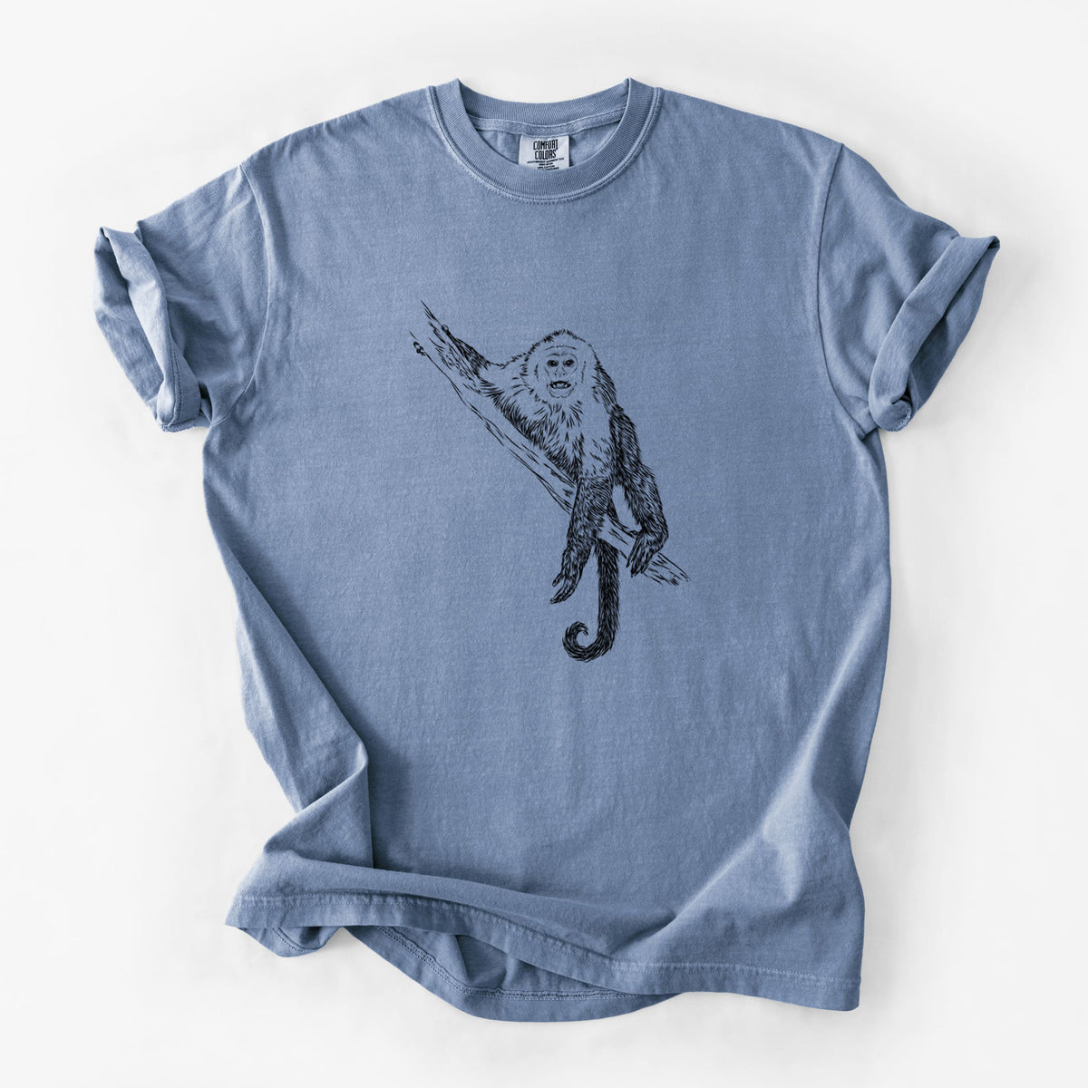Blue jean Comfort Colors t-shirt laid flat, printed with a detailed black ink illustration of a capuchin monkey climbing a branch.