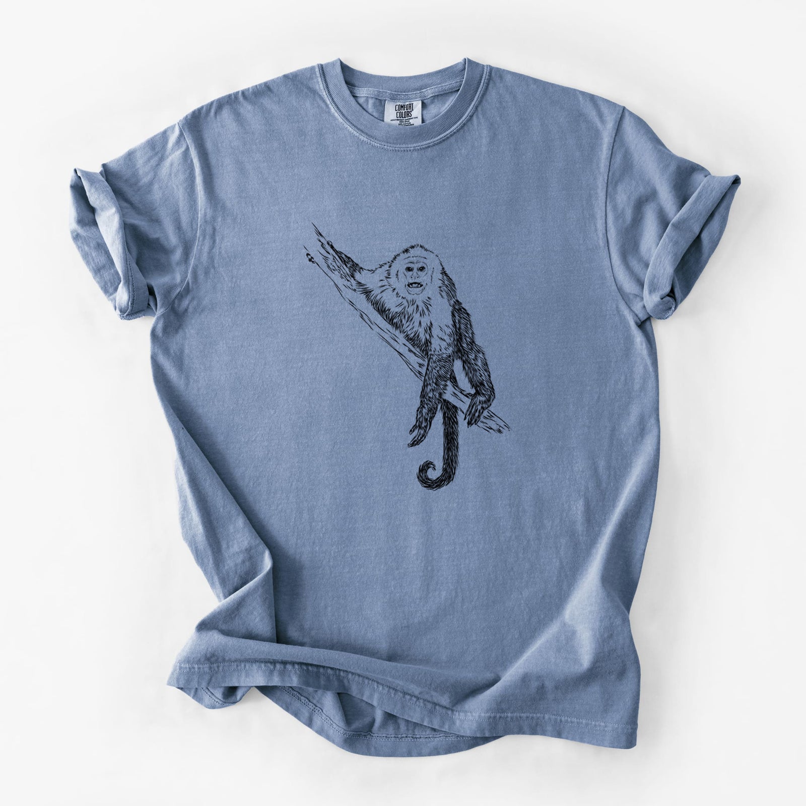 Blue jean Comfort Colors t-shirt laid flat, printed with a detailed black ink illustration of a capuchin monkey climbing a branch.