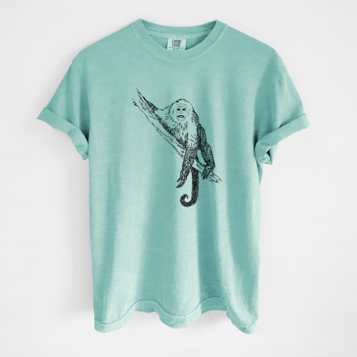 Chalky mint Comfort Colors t-shirt laid flat, printed with a detailed black ink illustration of a capuchin monkey climbing a branch.