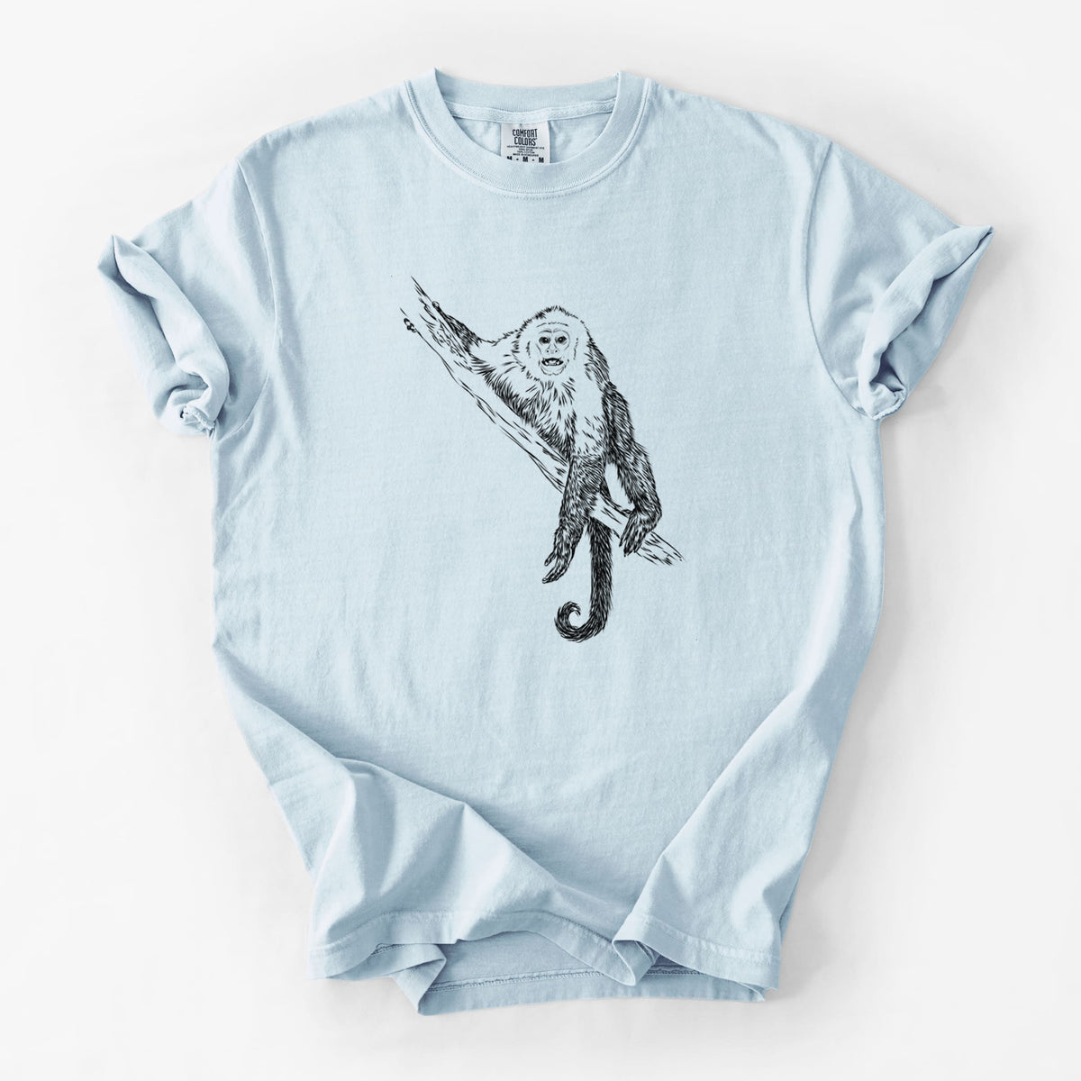 Chambray blue Comfort Colors t-shirt laid flat, printed with a detailed black ink illustration of a capuchin monkey climbing a branch.