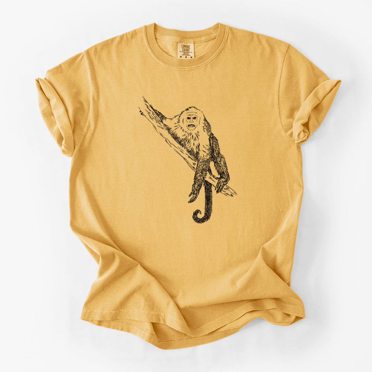 Golden wheat yellow Comfort Colors t-shirt laid flat, printed with a detailed black ink illustration of a capuchin monkey climbing a branch.