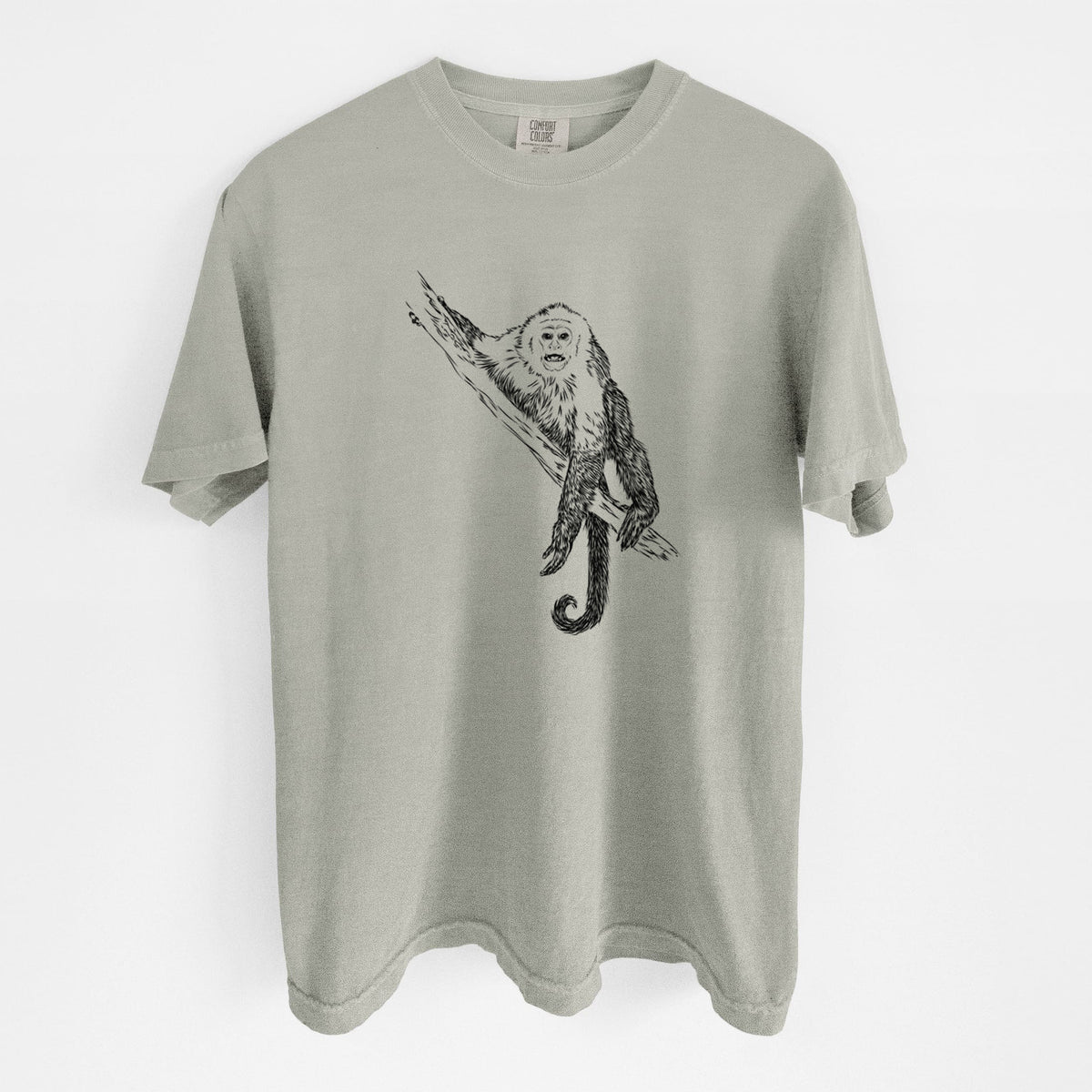 Sandstone beige Comfort Colors t-shirt laid flat, printed with a detailed black ink illustration of a capuchin monkey climbing a branch.