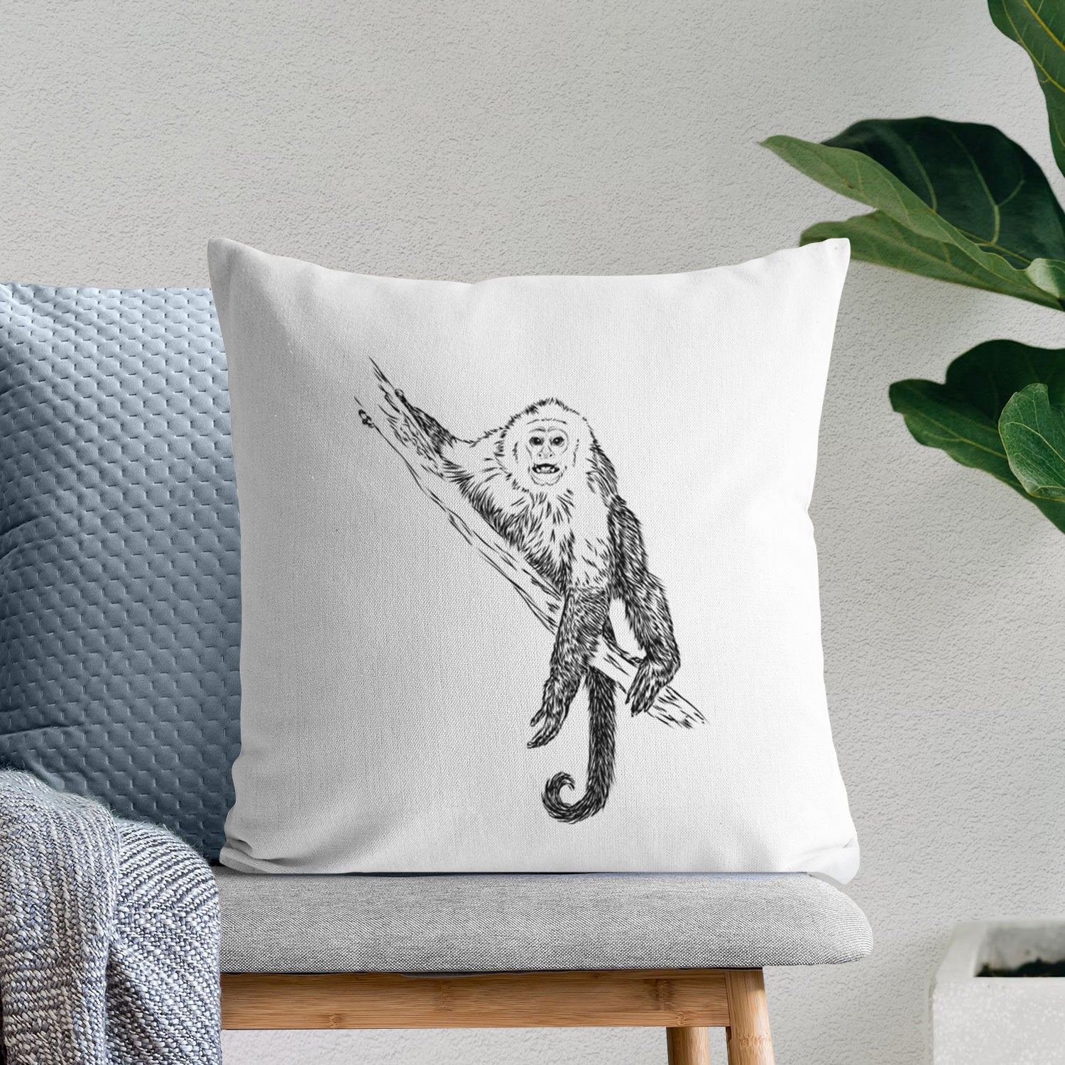 White pillow cover printed with a detailed black ink illustration of a capuchin monkey climbing a branch, on a bench beside plants.