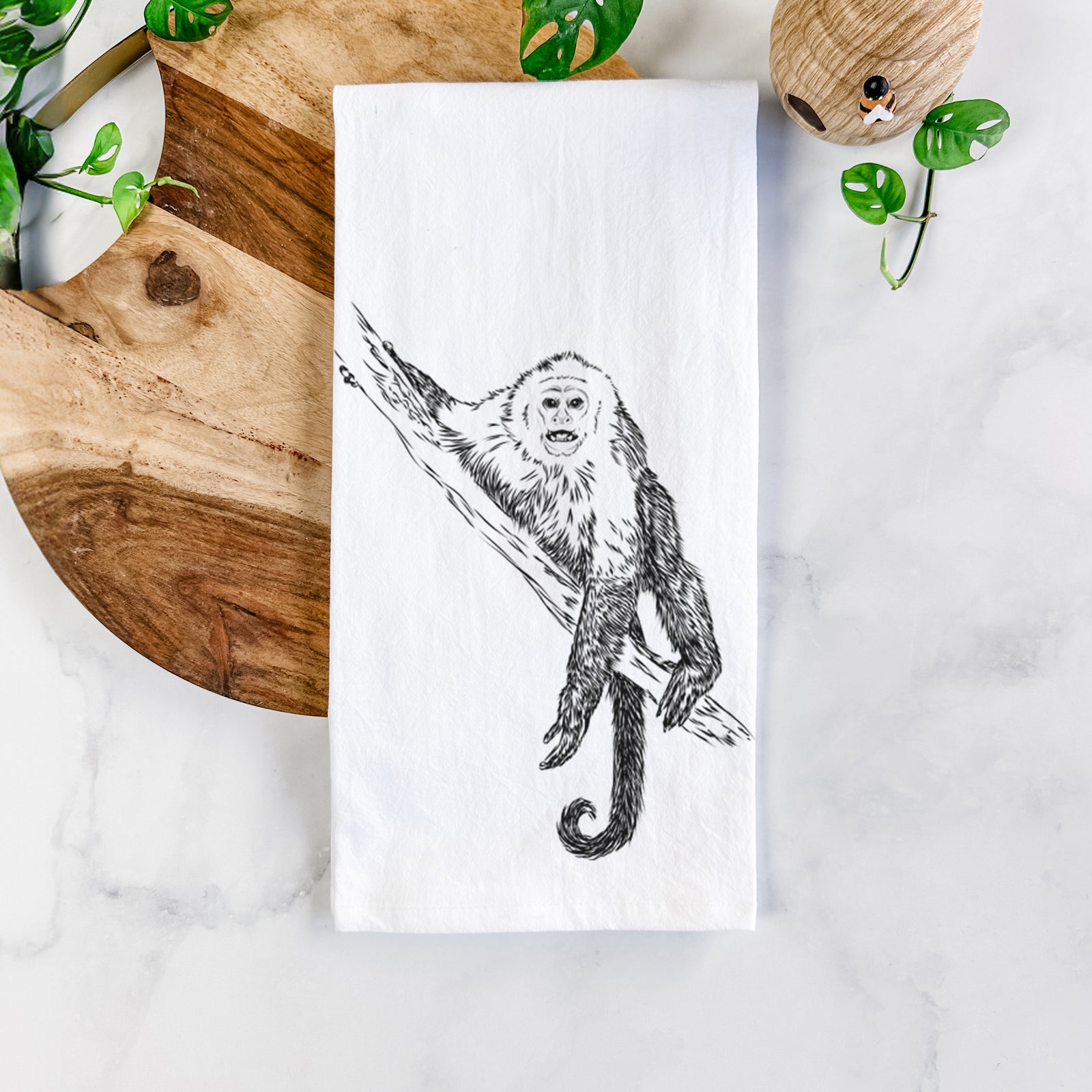 White cotton tea towel printed with a detailed black ink illustration of a capuchin monkey climbing a branch, on a marble counter beside a round wooden cutting board and greenery.