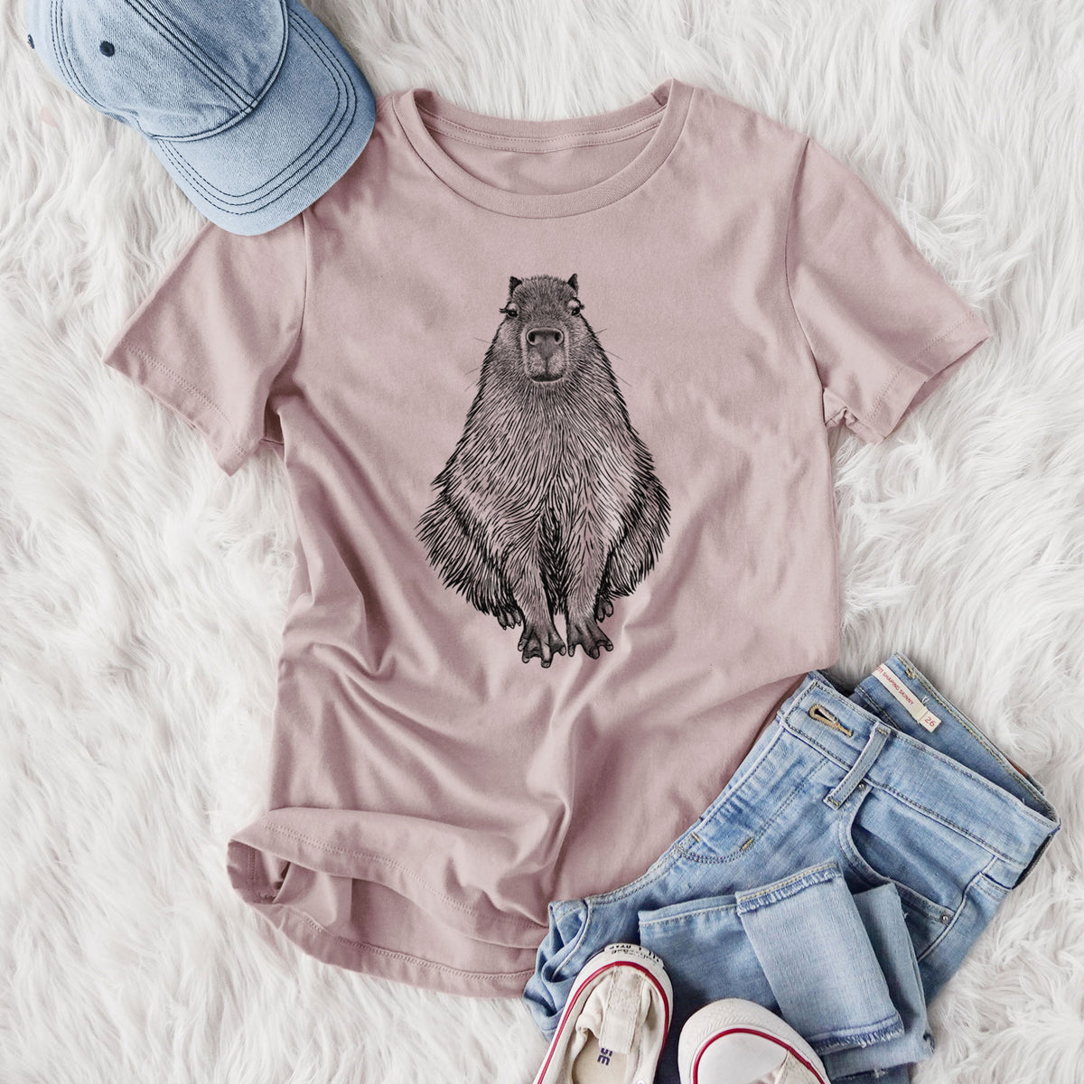 Women's relaxed fit cotton t-shirt printed with a detailed black ink illustration of a capybara sitting and facing forward, styled flat on a white faux fur rug with jeans.