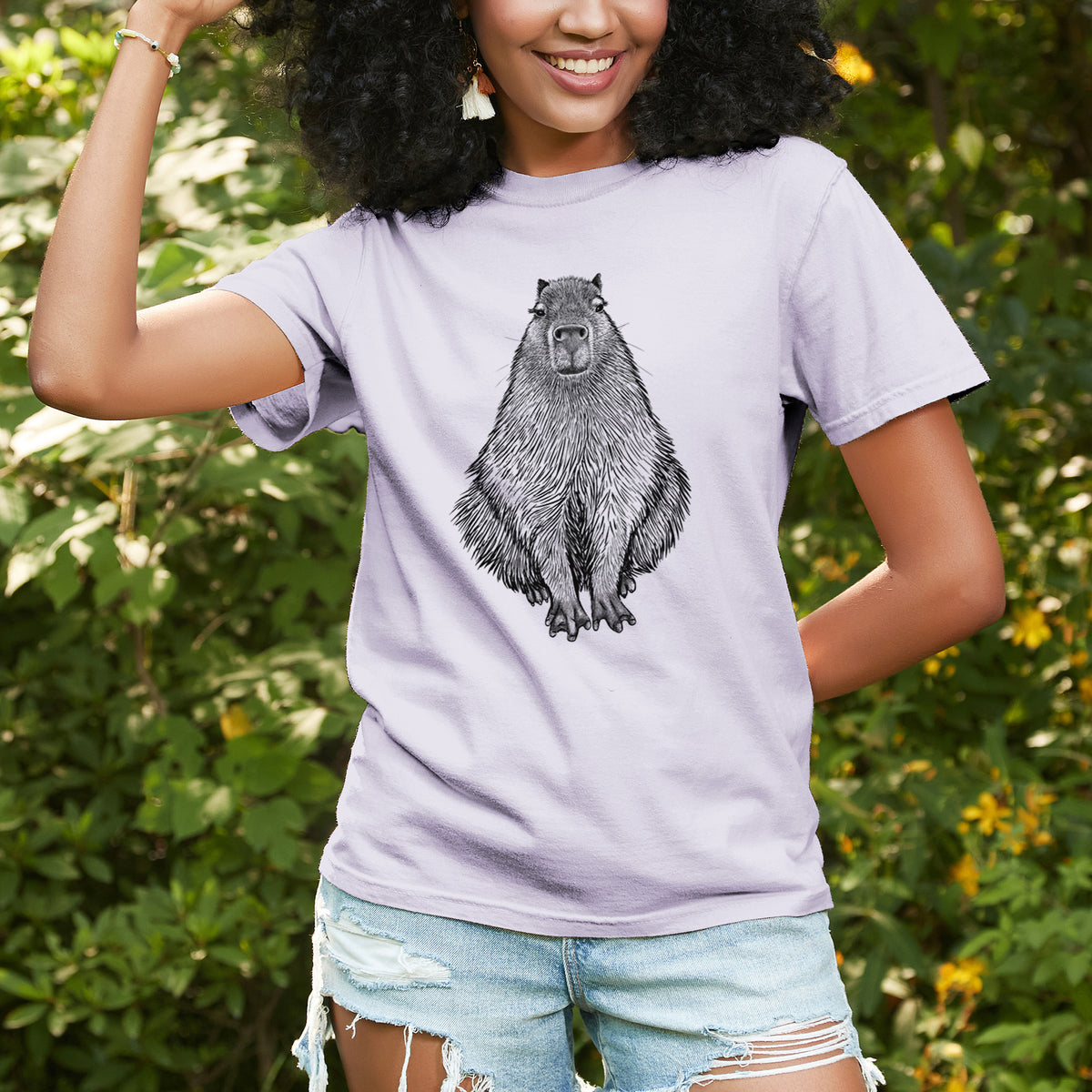 Woman wearing a Comfort Colors t-shirt printed with a detailed black ink illustration of a capybara sitting and facing forward, outdoors.
