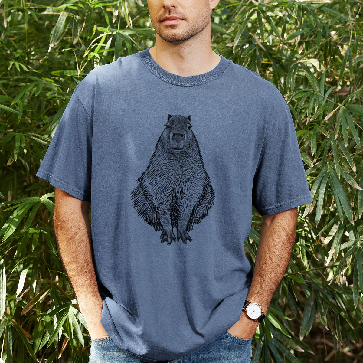 Man wearing a Comfort Colors t-shirt printed with a detailed black ink illustration of a capybara sitting and facing forward, outdoors.