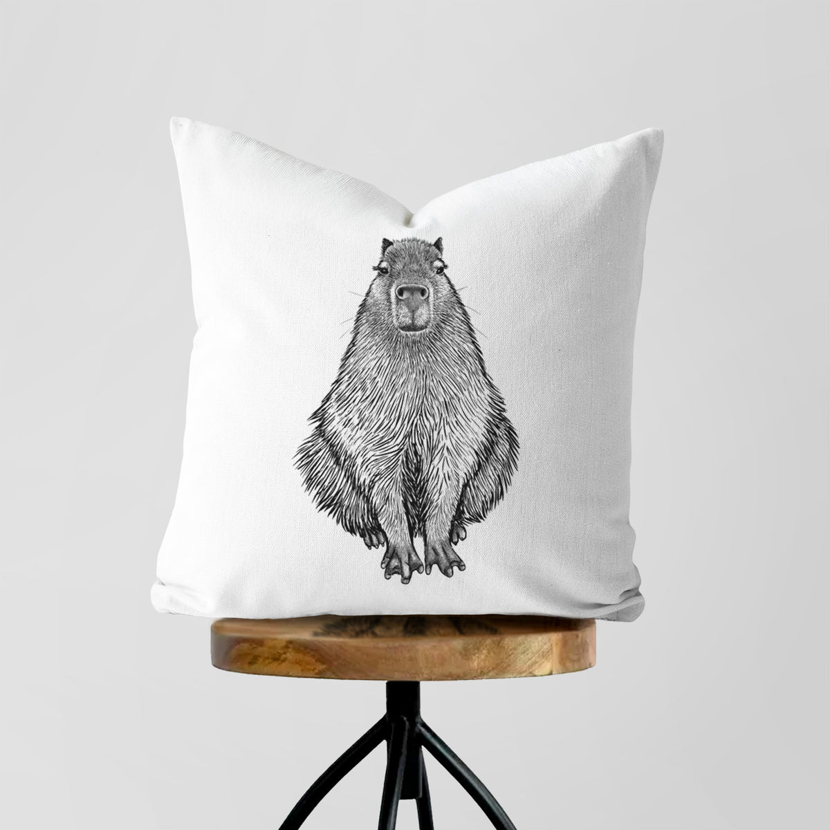 White pillow cover printed with a detailed black ink illustration of a capybara sitting and facing forward, on a wooden stool.