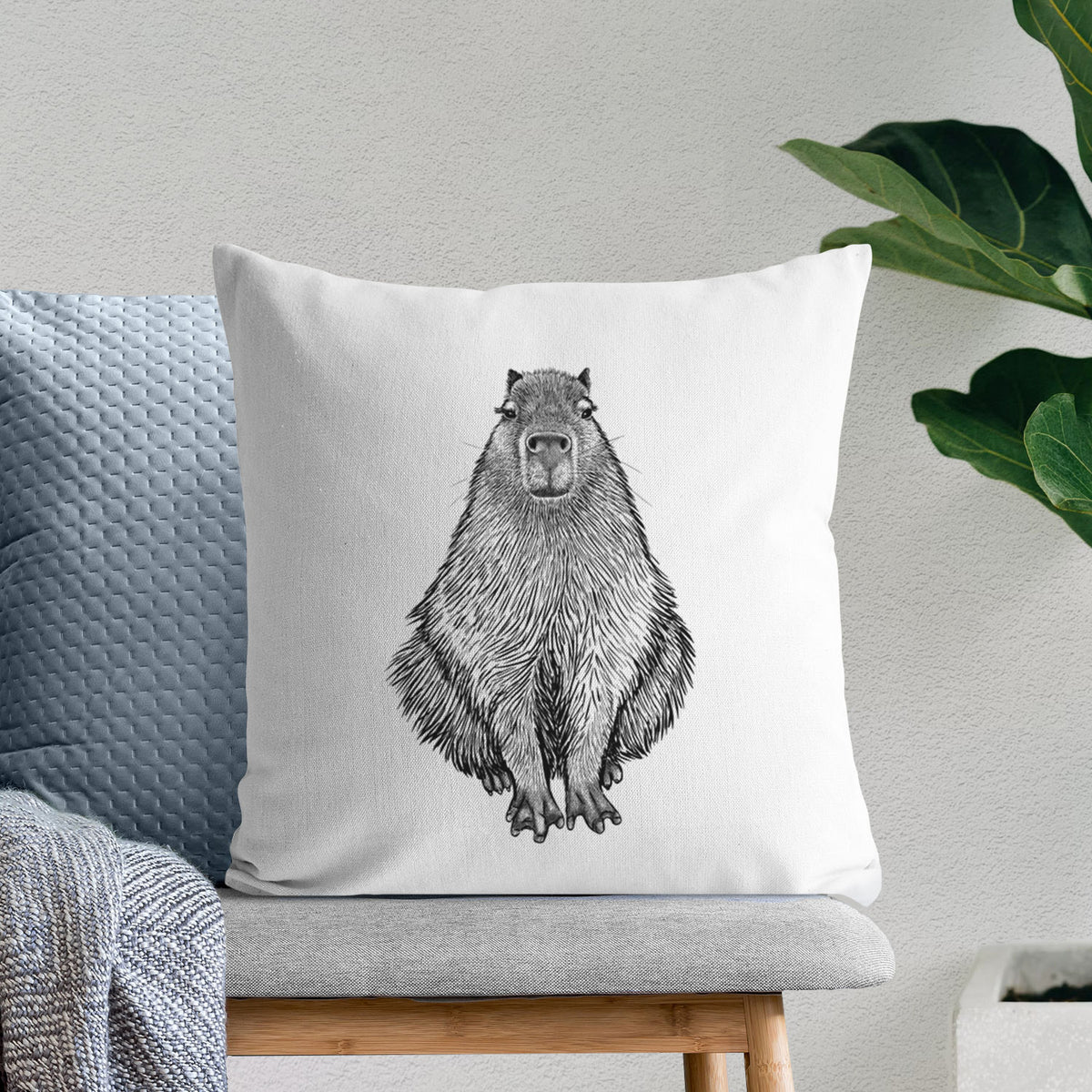 White pillow cover printed with a detailed black ink illustration of a capybara sitting and facing forward, on a bench beside plants.