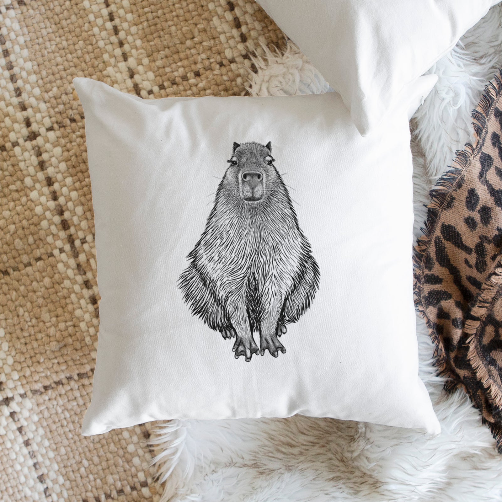 White pillow cover printed with a detailed black ink illustration of a capybara sitting and facing forward, on a bench beside plants.