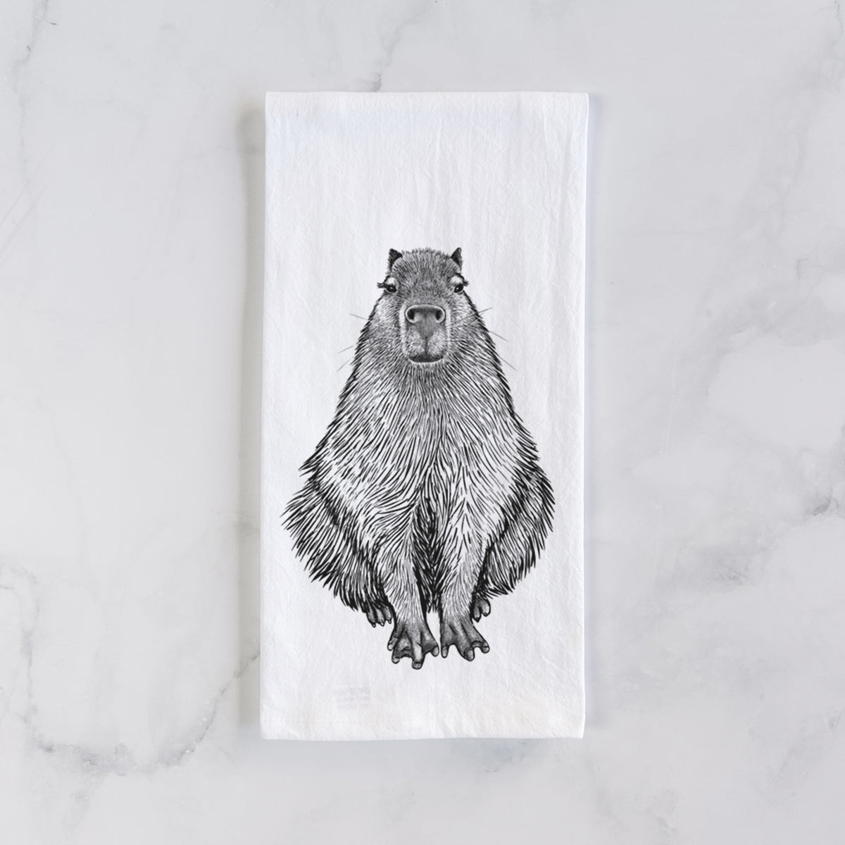 White cotton tea towel laid flat on marble, printed with a detailed black ink illustration of a capybara sitting and facing forward.