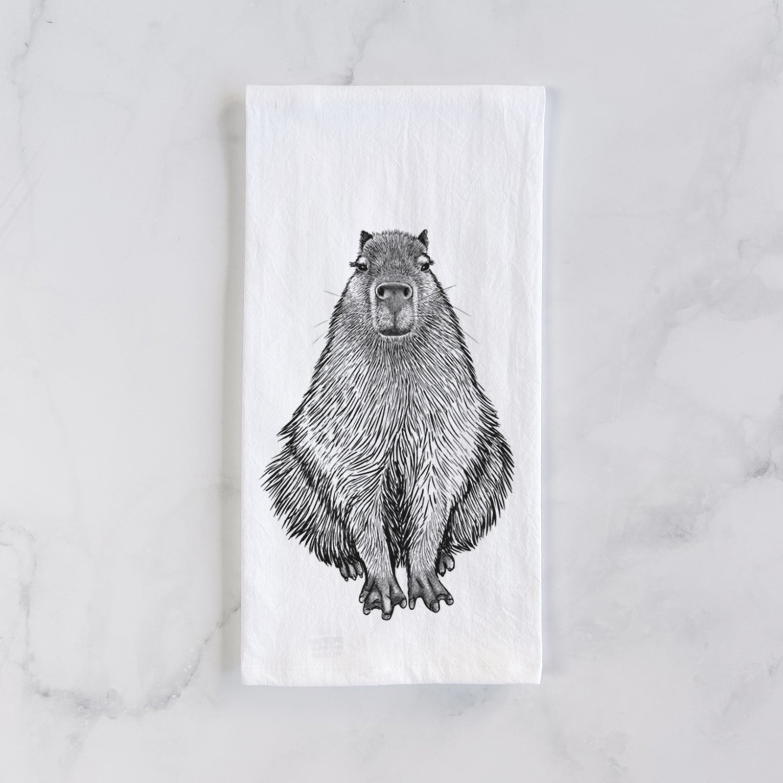 White cotton tea towel printed with a detailed black ink illustration of a capybara sitting and facing forward, on a marble counter beside a round wooden cutting board and greenery.