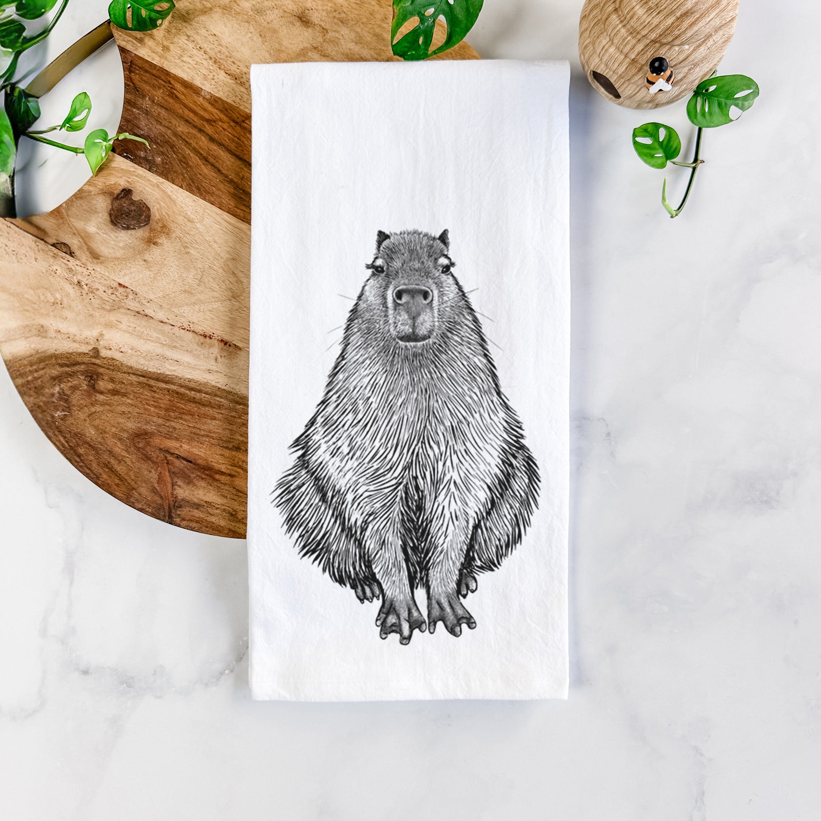 White cotton tea towel printed with a detailed black ink illustration of a capybara sitting and facing forward, on a marble counter beside a round wooden cutting board and greenery.
