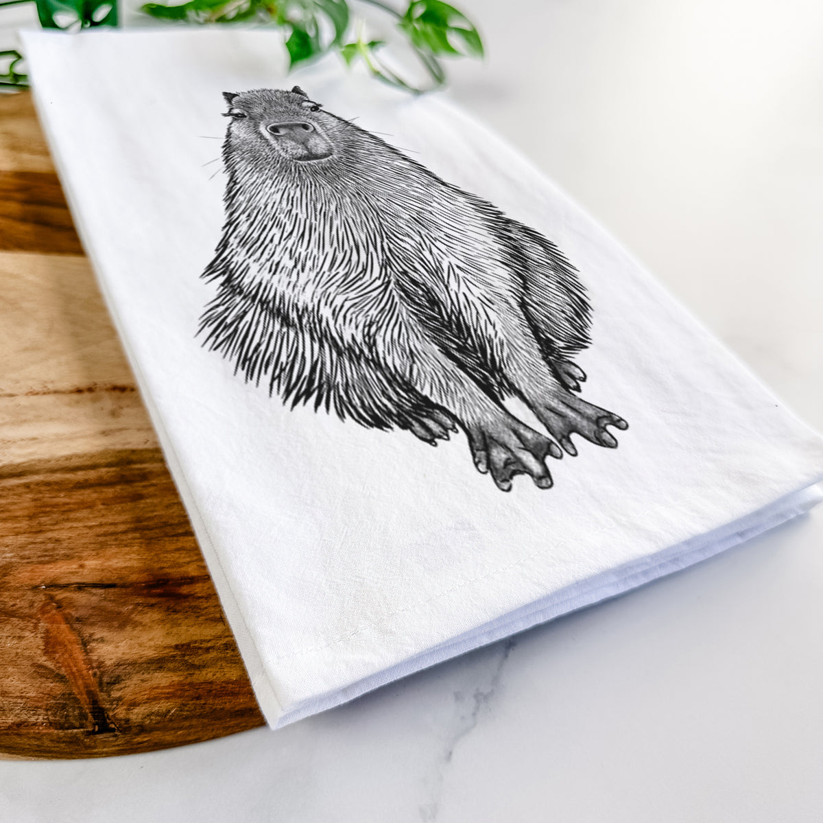 Close-up of a folded white tea towel on a wooden board, showing a detailed black ink illustration of a capybara sitting and facing forward.