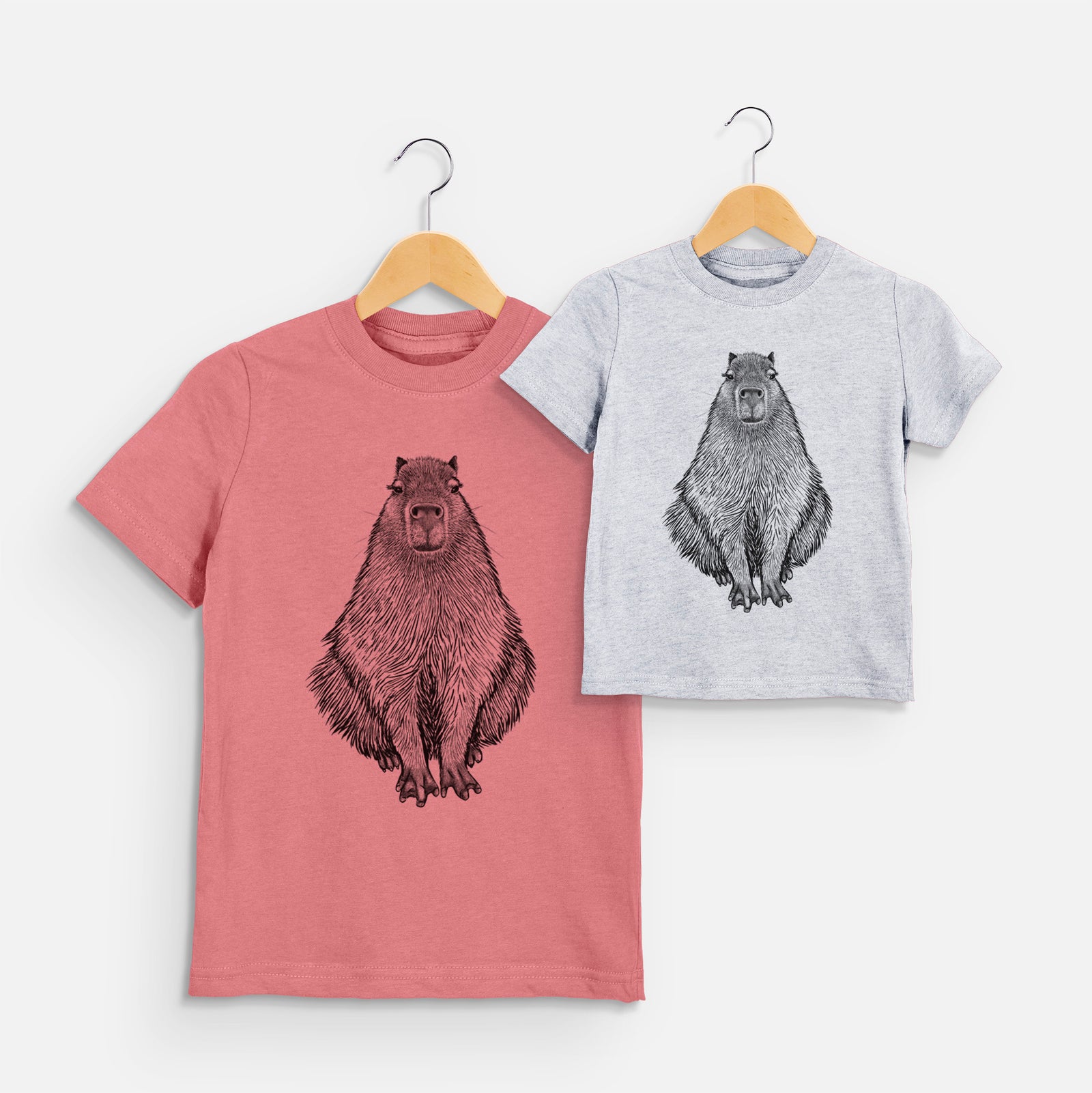 Two youth t-shirts in different colors on hangers, printed with a detailed black ink illustration of a capybara sitting and facing forward.