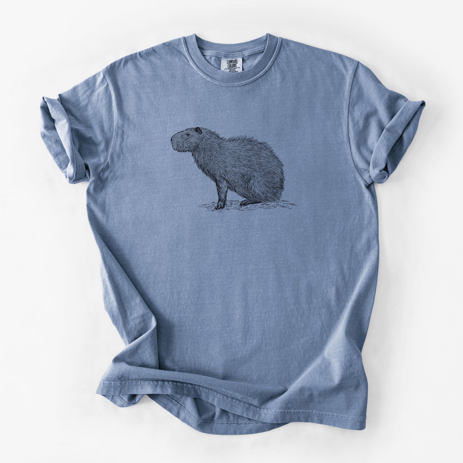 Blue jean Comfort Colors t-shirt laid flat, printed with a detailed black ink illustration of a capybara standing in profile.