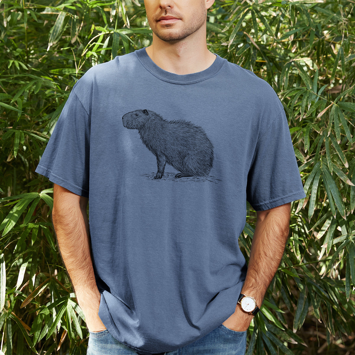Man wearing a Comfort Colors t-shirt printed with a detailed black ink illustration of a capybara standing in profile, outdoors.