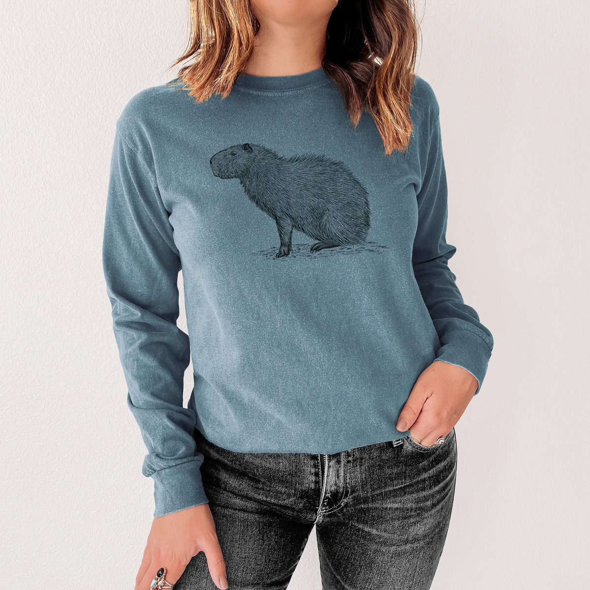 Steel blue Comfort Colors long sleeve shirt printed with a detailed black ink illustration of a capybara standing in profile, shown in a lifestyle photo.