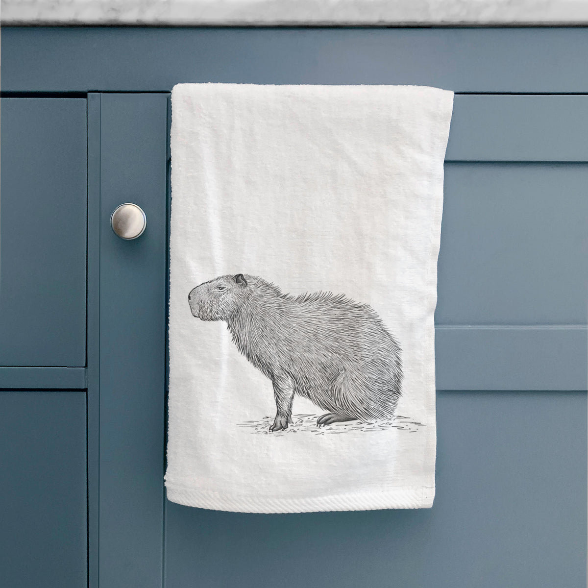 White hand towel printed with a detailed black ink illustration of a capybara standing in profile, hanging on a cabinet door.